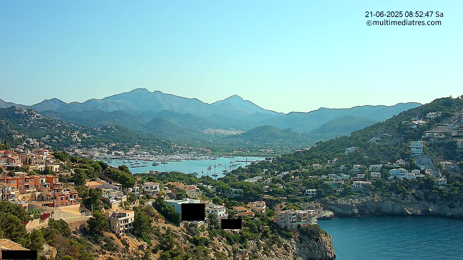 A coastal town nestled against a mountain range under a clear blue sky, with a calm bay filled with sailboats in the foreground.