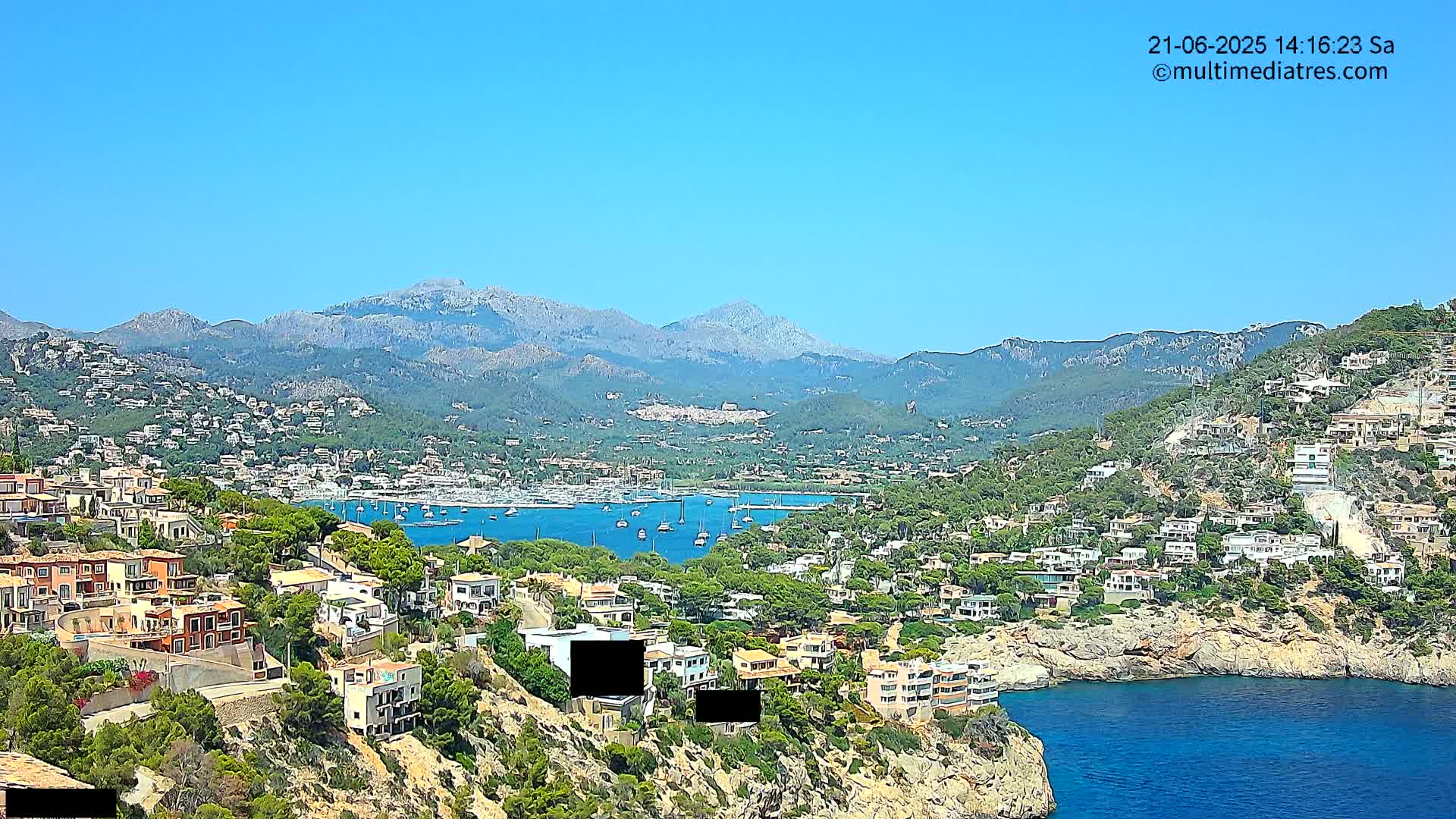 A coastal town nestled against a mountain range on a clear, sunny day, with a harbor filled with boats in the foreground.