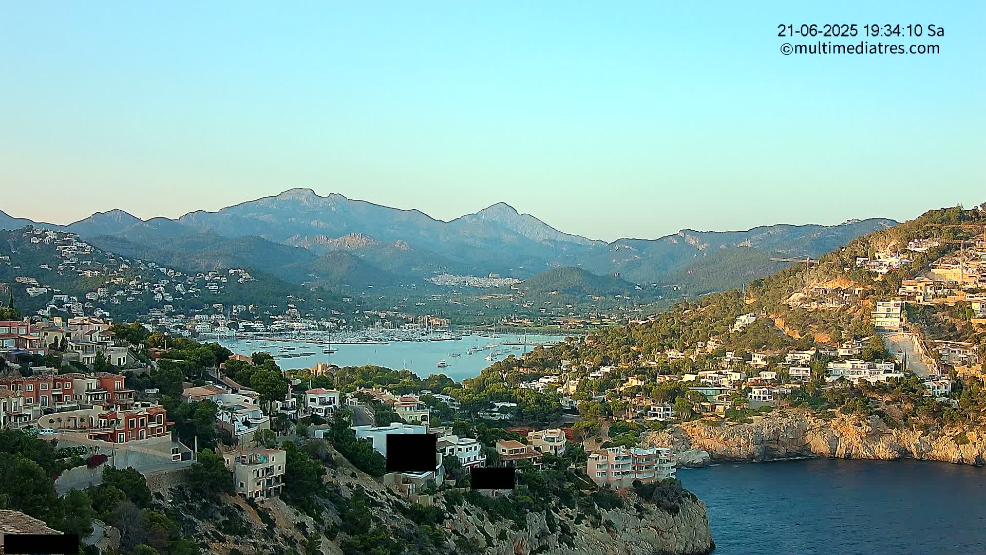 A coastal town nestled between a calm bay filled with boats and a mountain range under a clear evening sky.