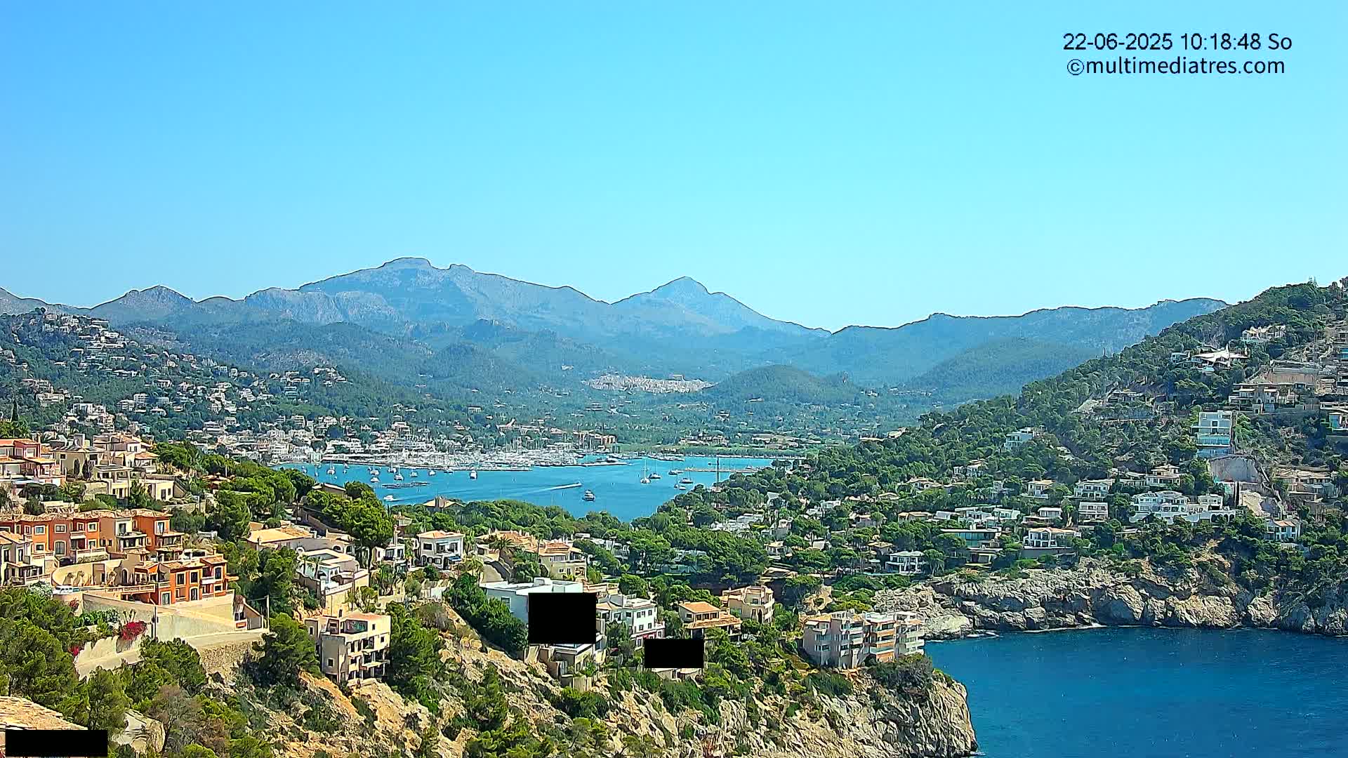 A coastal town nestled against a mountain range under a clear blue sky, with a calm body of water filled with sailboats.