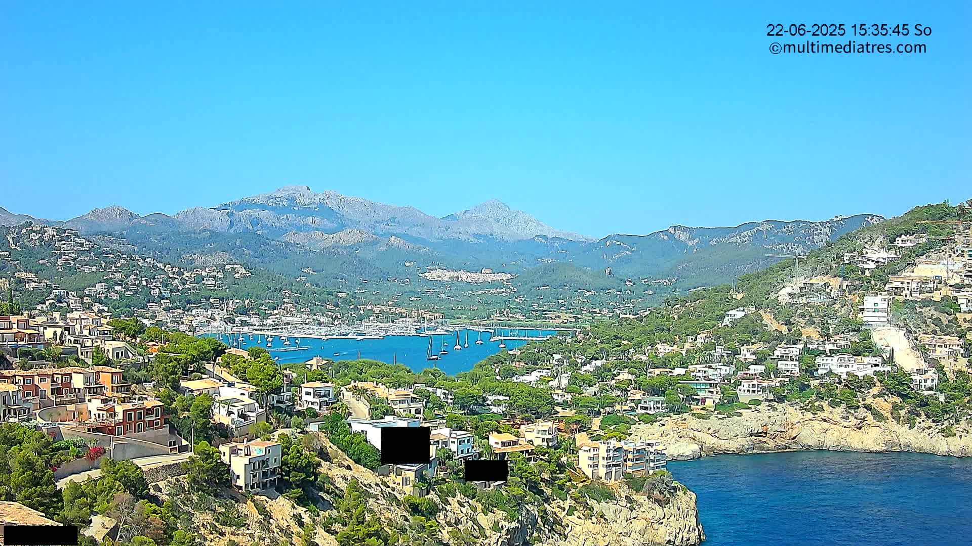 A coastal town nestled against a mountain range under a clear blue sky, with a harbor full of sailboats visible in the distance.