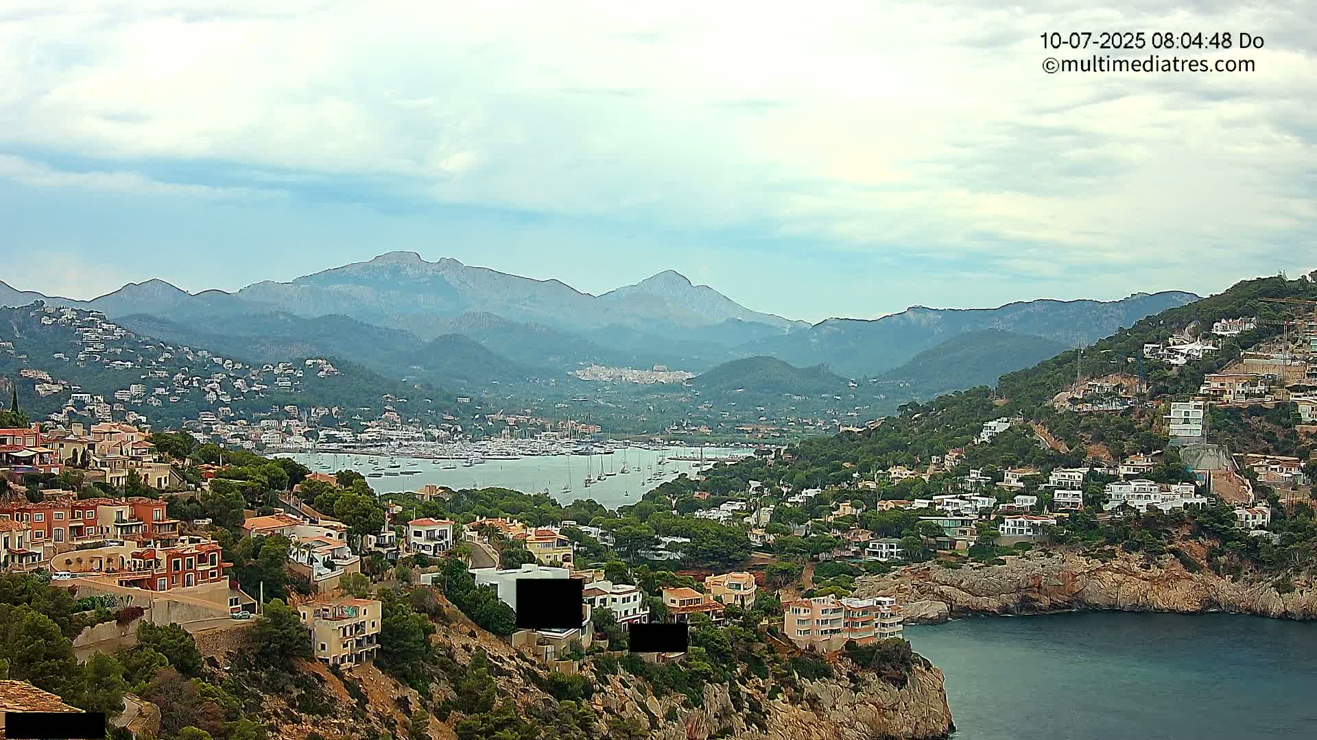 A coastal town nestled among hills and mountains overlooks a calm bay filled with sailboats under a partly cloudy sky.