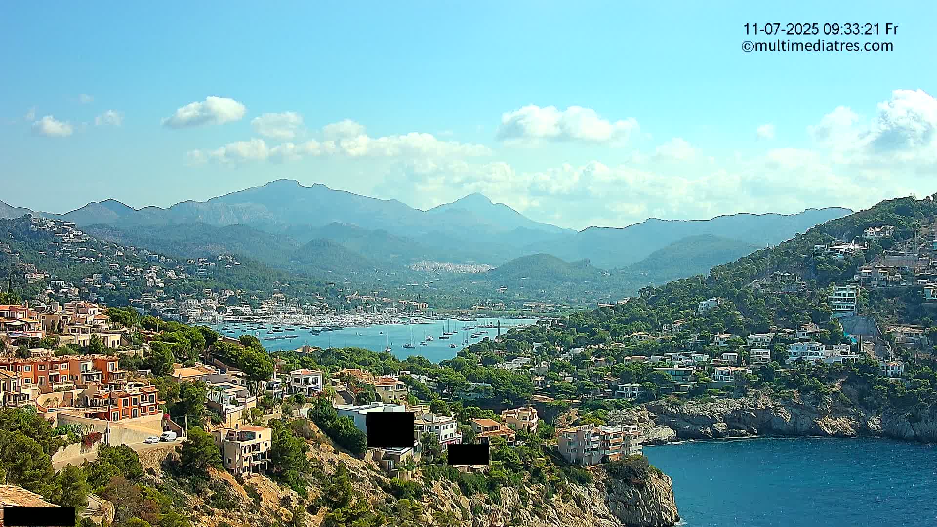 A coastal town nestled among hills and mountains under a mostly clear sky, with a calm bay filled with numerous boats.