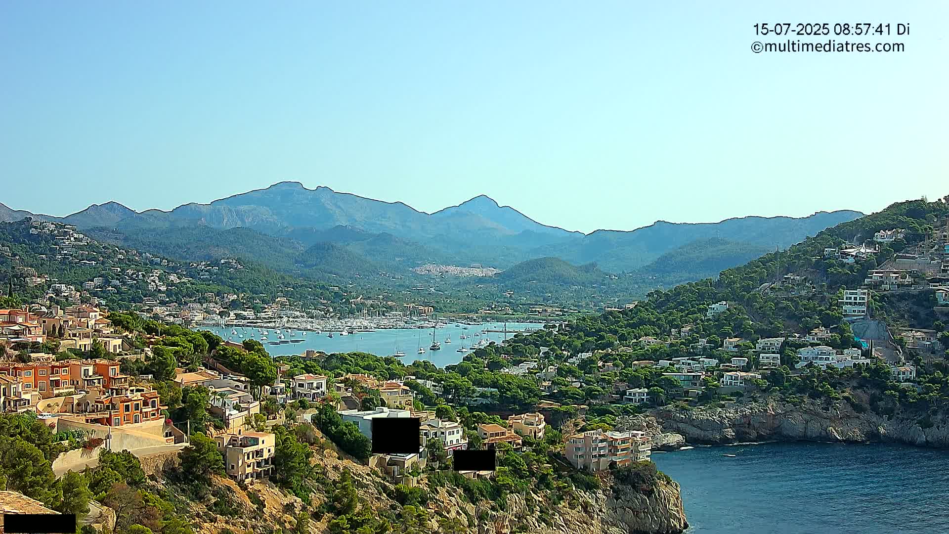A coastal town nestled against a mountain range under a clear blue sky overlooks a harbor filled with boats.