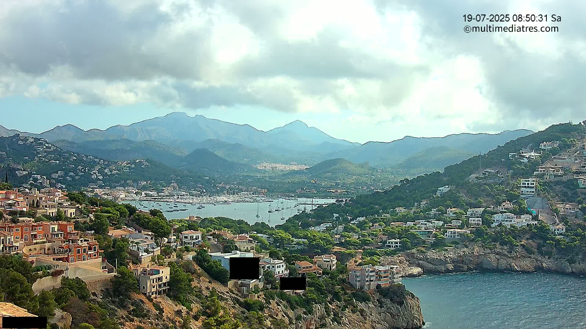 A coastal town nestled on a hillside overlooking a harbor filled with sailboats, with mountains in the background under a partly cloudy sky.