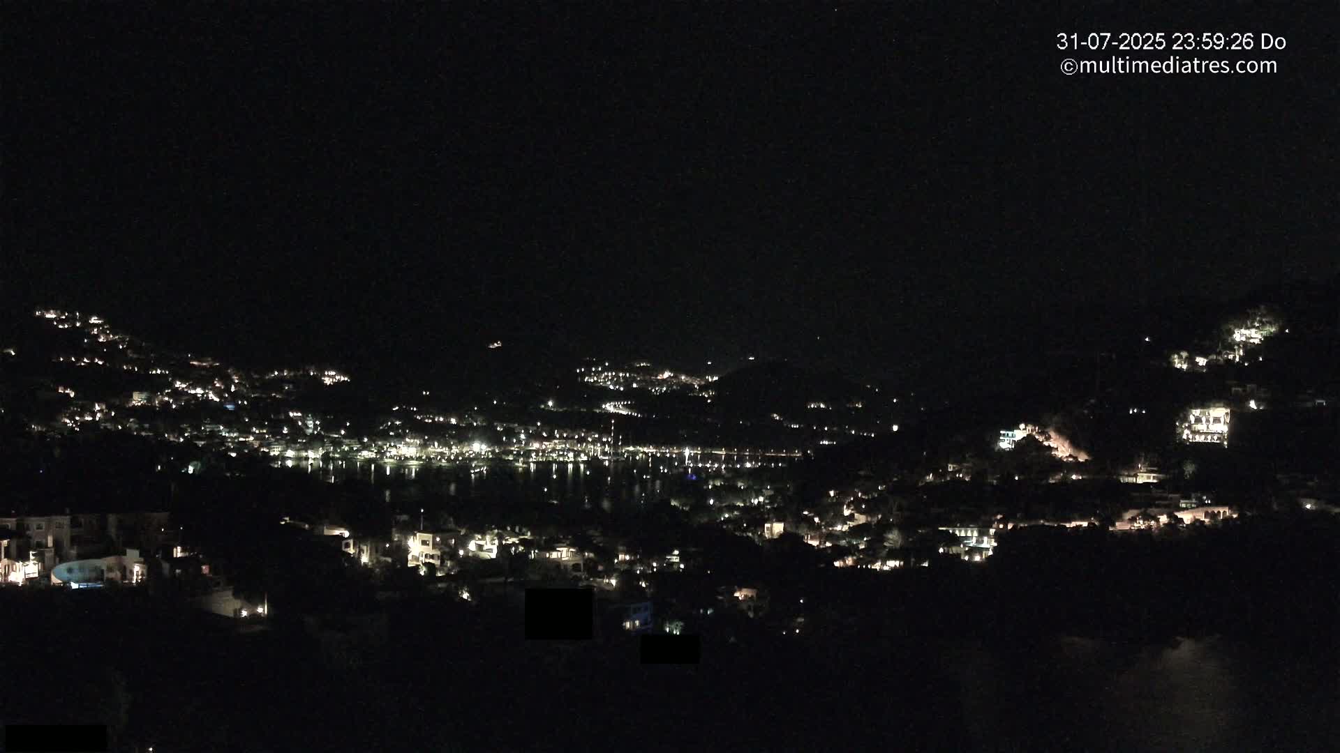 A nighttime view of a coastal town nestled on hillsides, with numerous lights illuminating houses and a dark body of water below.