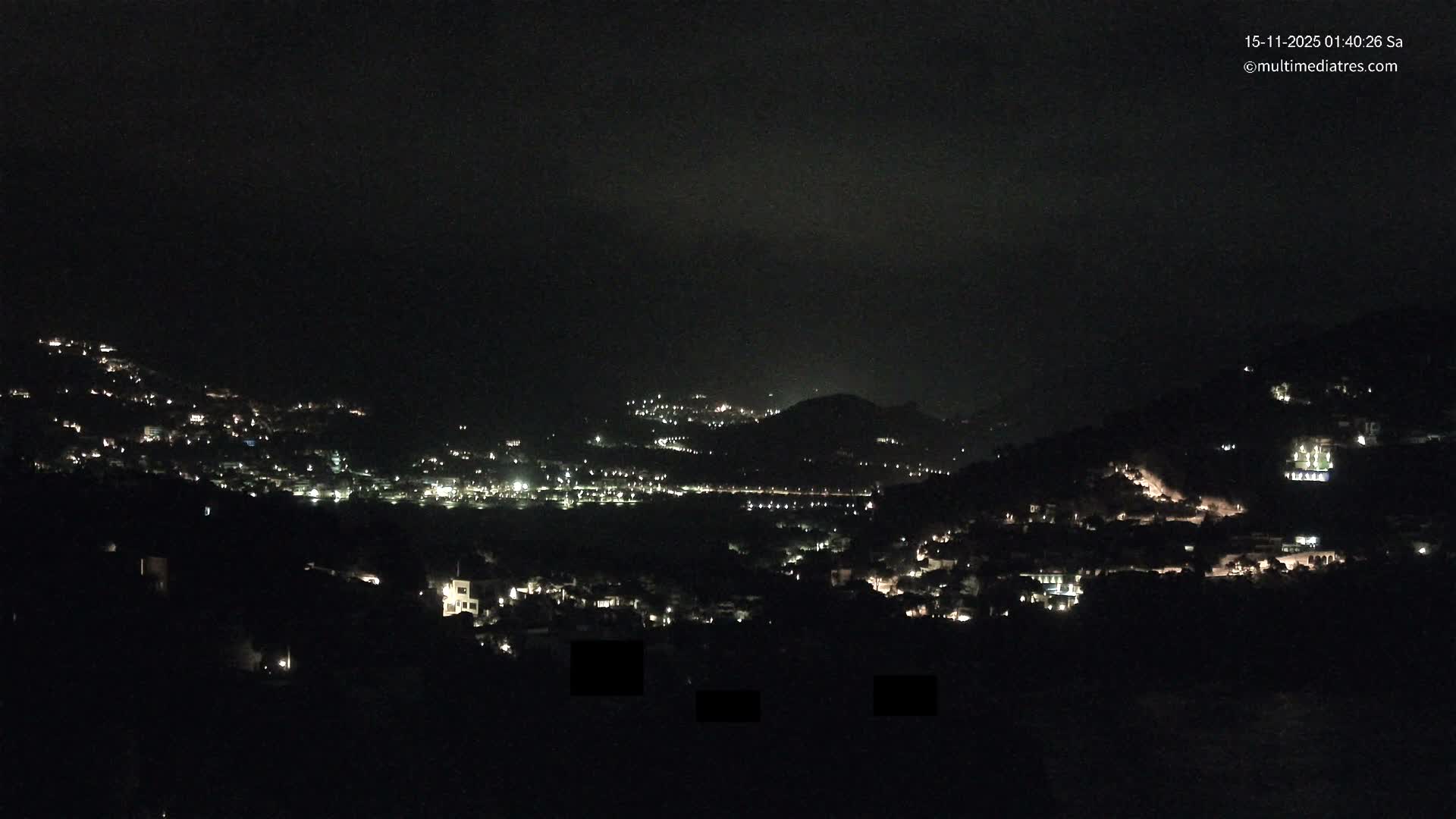 A panoramic nighttime view reveals the twinkling lights of a town spread across hills and valleys under a dark, slightly hazy sky.