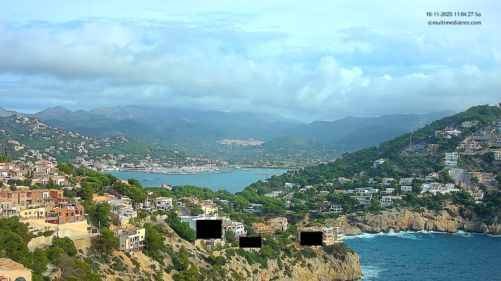 A picturesque coastal town spreads across verdant hillsides and rocky cliffs, overlooking a tranquil bay dotted with boats, framed by distant mountains, all beneath a partly cloudy sky with light waves breaking on the shore.