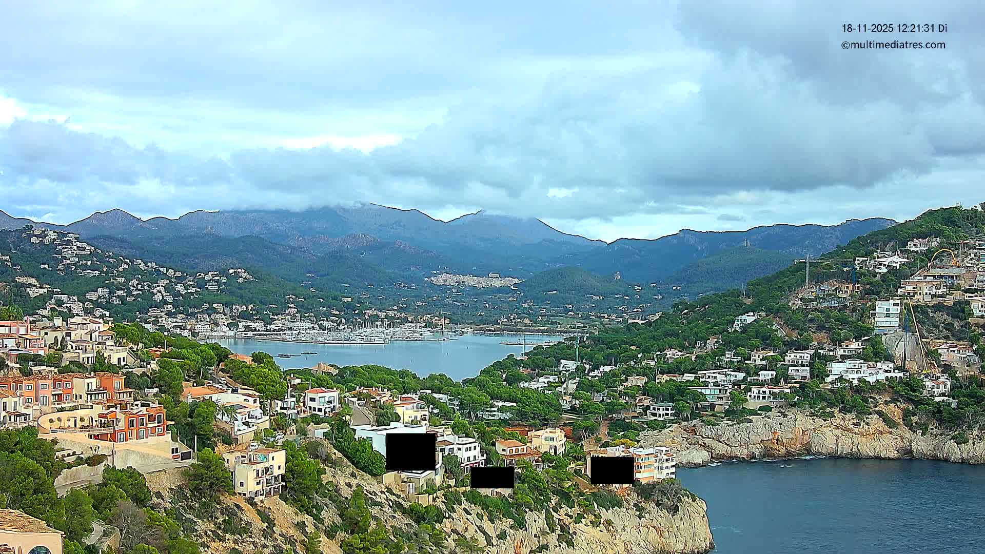 A picturesque coastal town spreads across verdant hillsides and rocky cliffs, overlooking a tranquil bay dotted with boats, framed by distant mountains, all beneath a partly cloudy sky with light waves breaking on the shore.