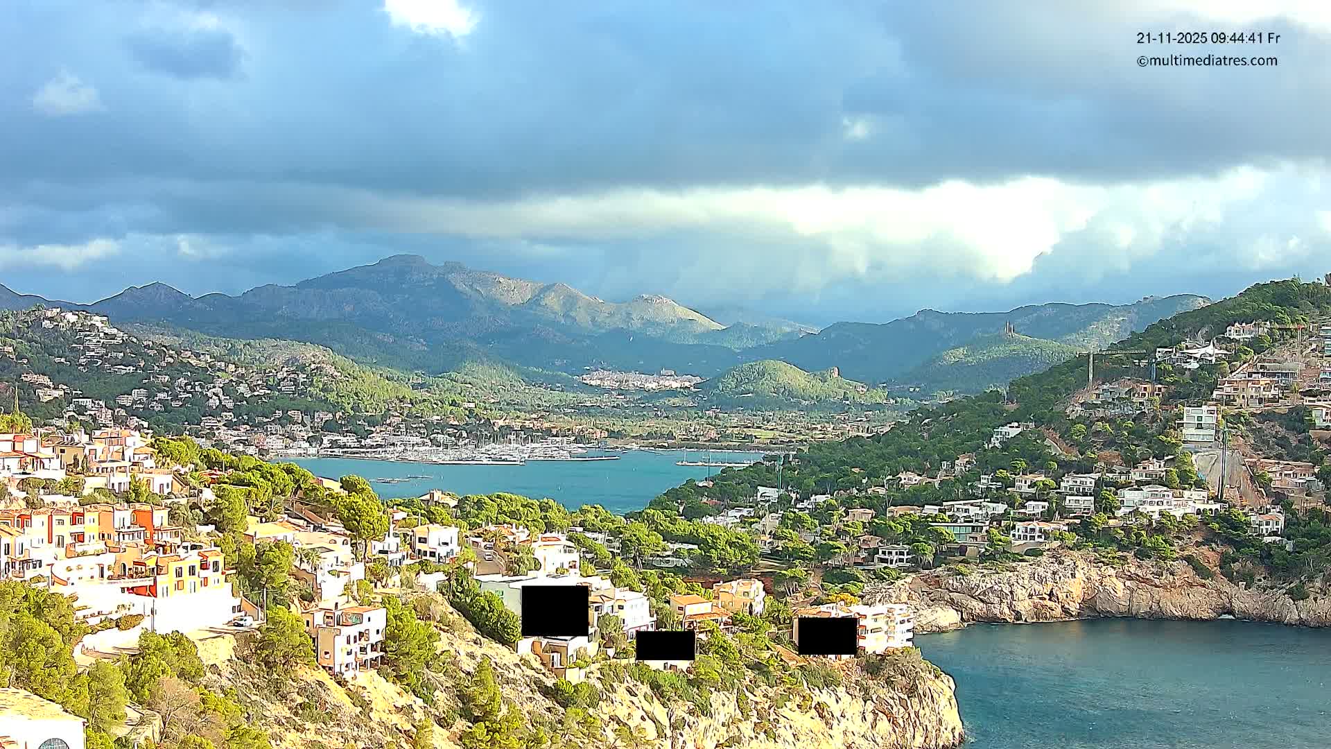 A picturesque coastal town with many white and terracotta buildings covers lush green hillsides overlooking a serene blue bay dotted with boats, all set against a backdrop of distant mountains beneath a partly cloudy sky where sunlight breaks through.