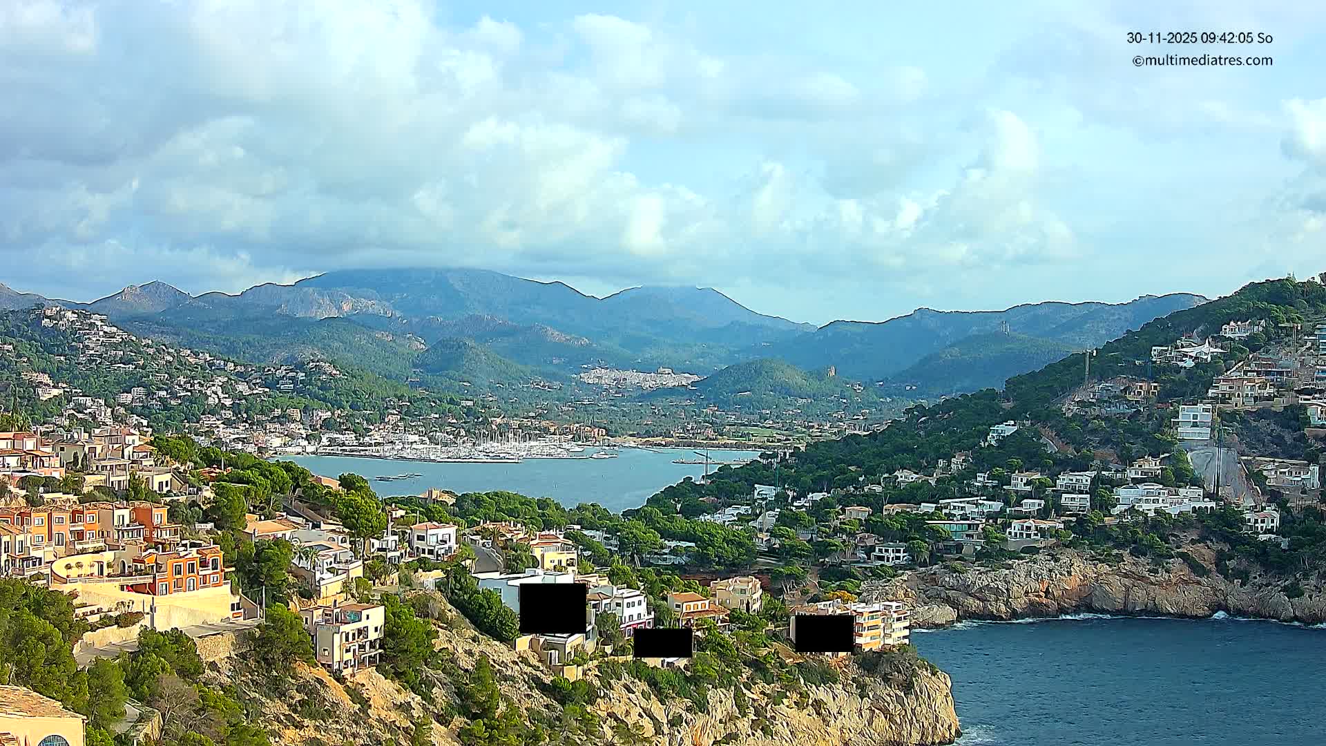 A scenic coastal town with houses sprawling across tree-covered hillsides overlooks a large bay dotted with boats and rocky cliffs, all under a partly cloudy sky.