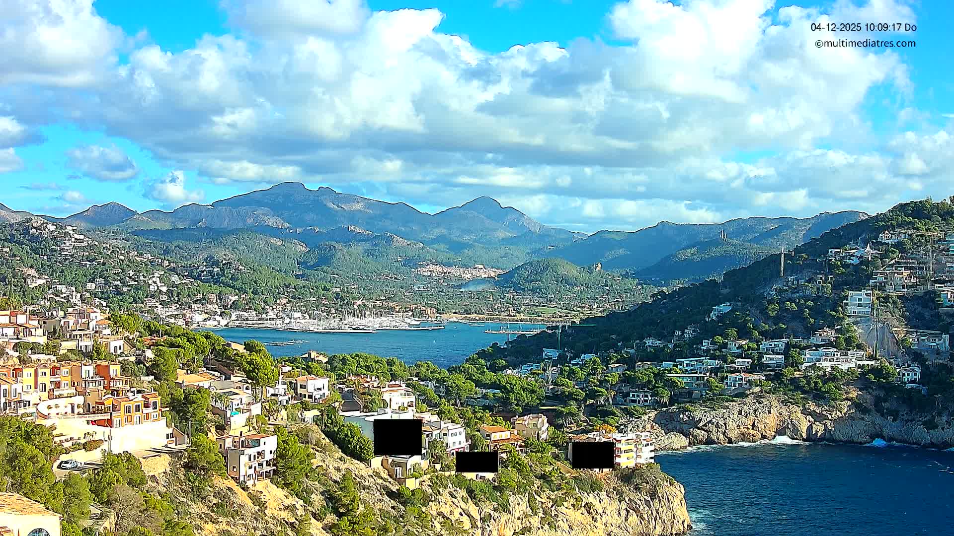 An expansive outdoor shot reveals a vibrant coastal town nestled into lush hillsides overlooking a serene bay filled with boats and backed by majestic mountains, all under a bright, partly cloudy sky.