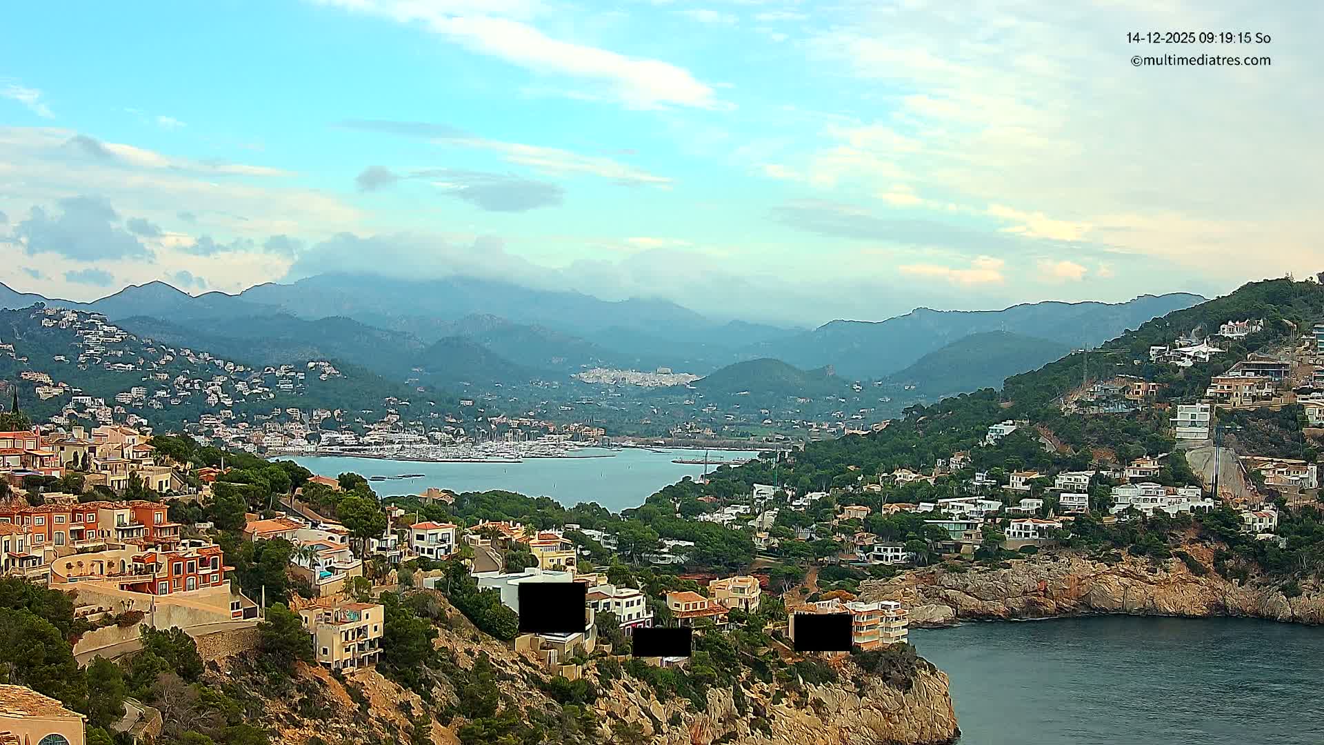 An expansive outdoor shot reveals a vibrant coastal town nestled into lush hillsides overlooking a serene bay filled with boats and backed by majestic mountains, all under a bright, partly cloudy sky.