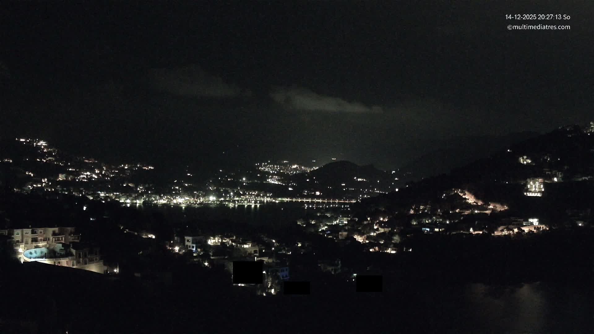 An expansive outdoor shot reveals a vibrant coastal town nestled into lush hillsides overlooking a serene bay filled with boats and backed by majestic mountains, all under a bright, partly cloudy sky.