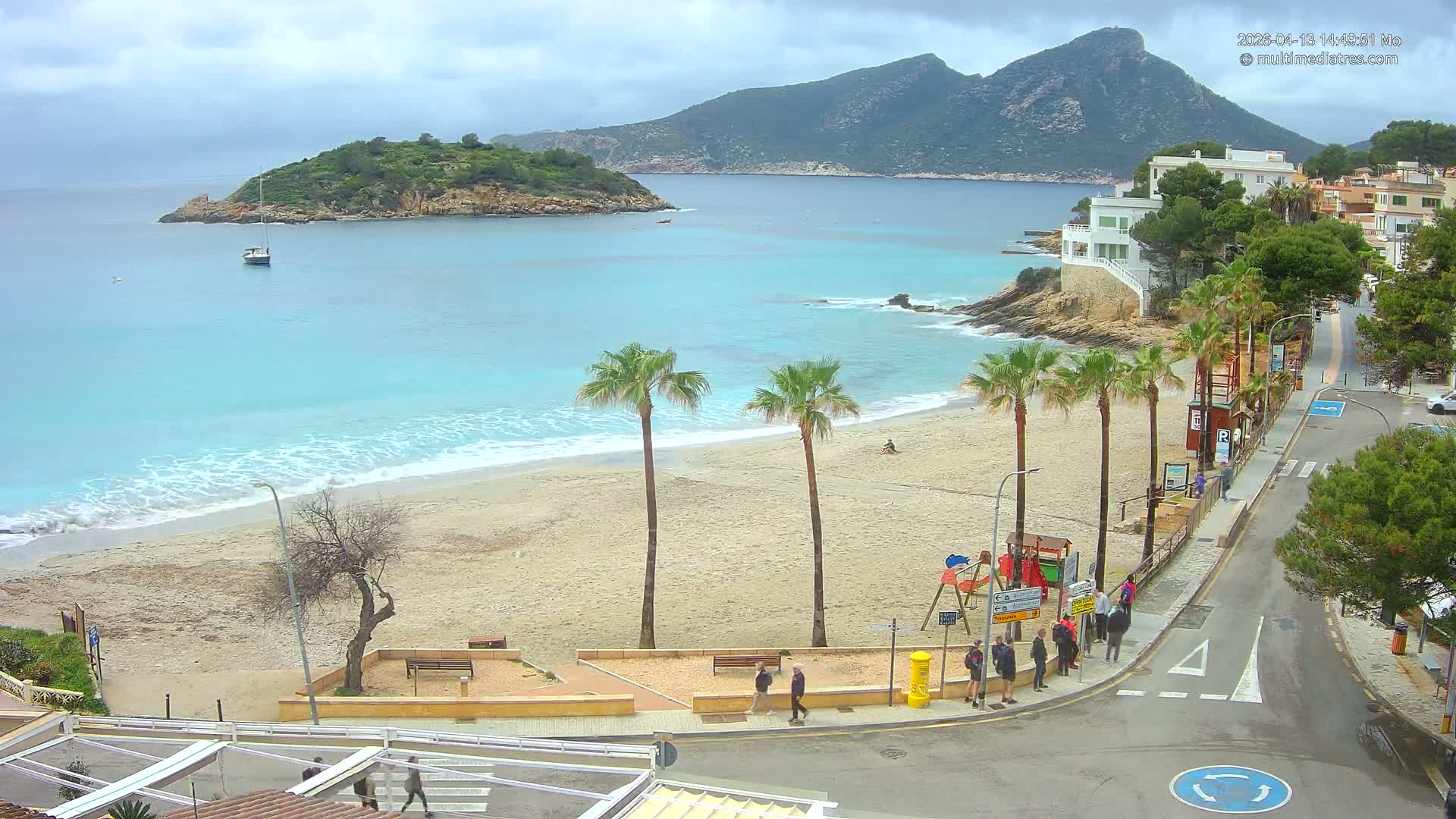 An expansive outdoor shot reveals a vibrant coastal town nestled into lush hillsides overlooking a serene bay filled with boats and backed by majestic mountains, all under a bright, partly cloudy sky.