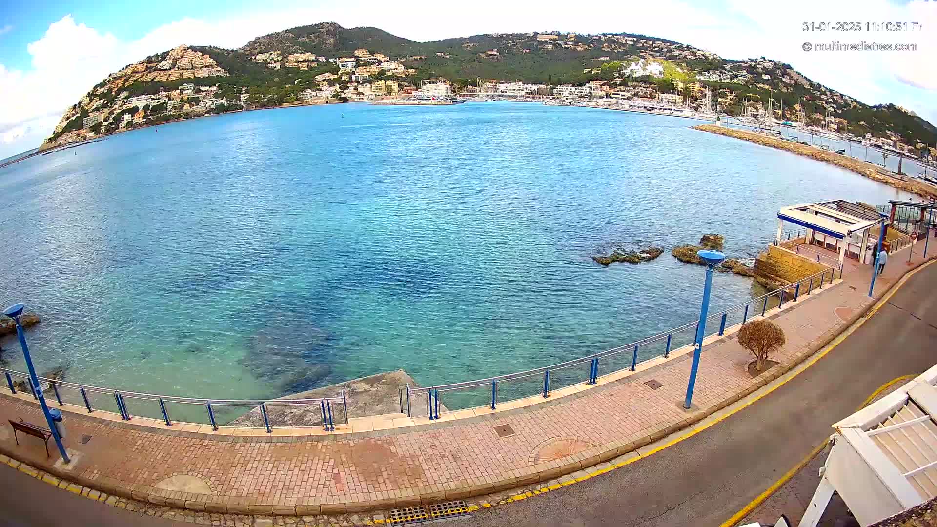 A partly sunny day shows a seaside town nestled against a hill overlooking a calm, clear blue bay with a paved walkway and a small structure along the shore.