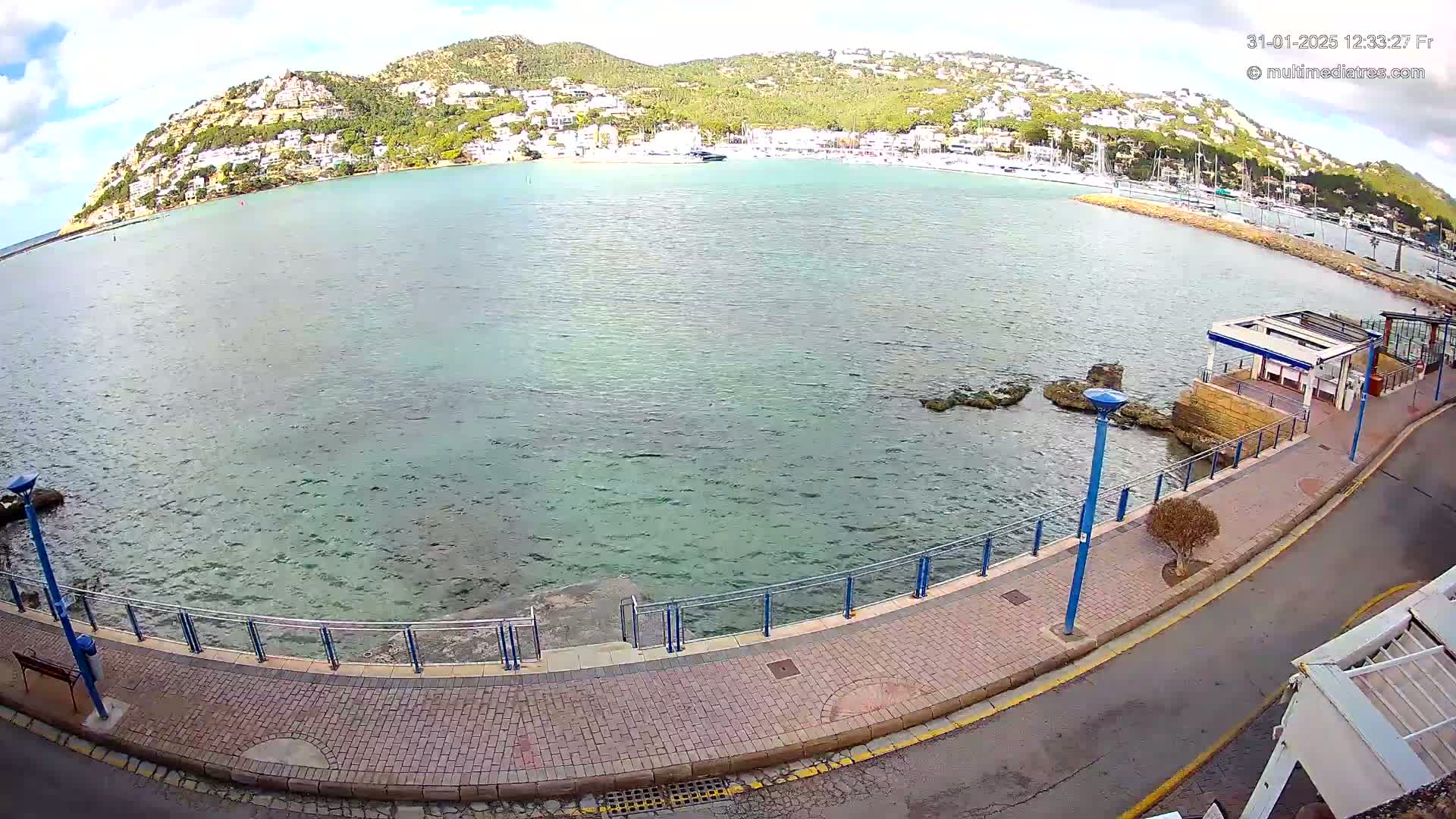 A waterfront paved walkway with a metal railing overlooks a calm, greenish-blue sea, with a hillside town visible in the background under a partly cloudy sky.
