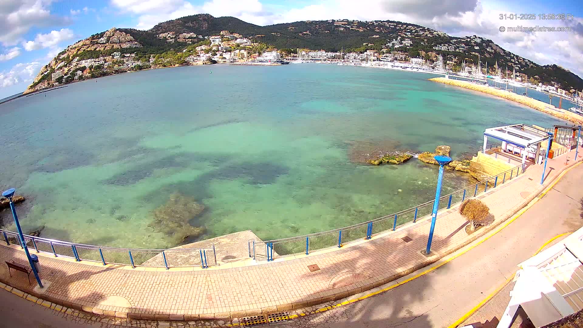 A clear, sunny day shows a paved walkway next to a calm, shallow, turquoise body of water with a hill covered in buildings in the background.