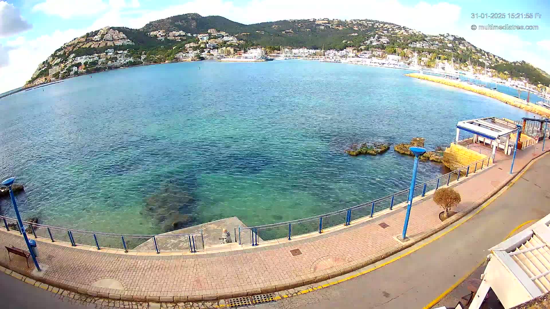 A partly sunny day overlooks a calm, clear blue bay with a town and hills in the background, as seen from a paved walkway with a railing overlooking the water.