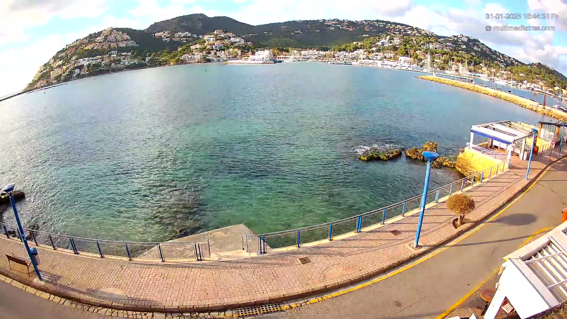 A paved walkway curves along a calm, clear-water bay, overlooking a hillside town under a partly cloudy sky.