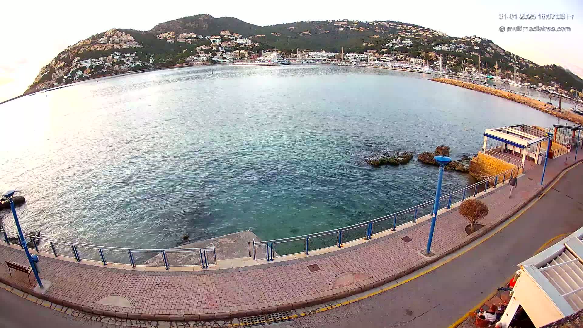 A calm bay with a town nestled on a hillside overlooking a paved walkway next to the water.