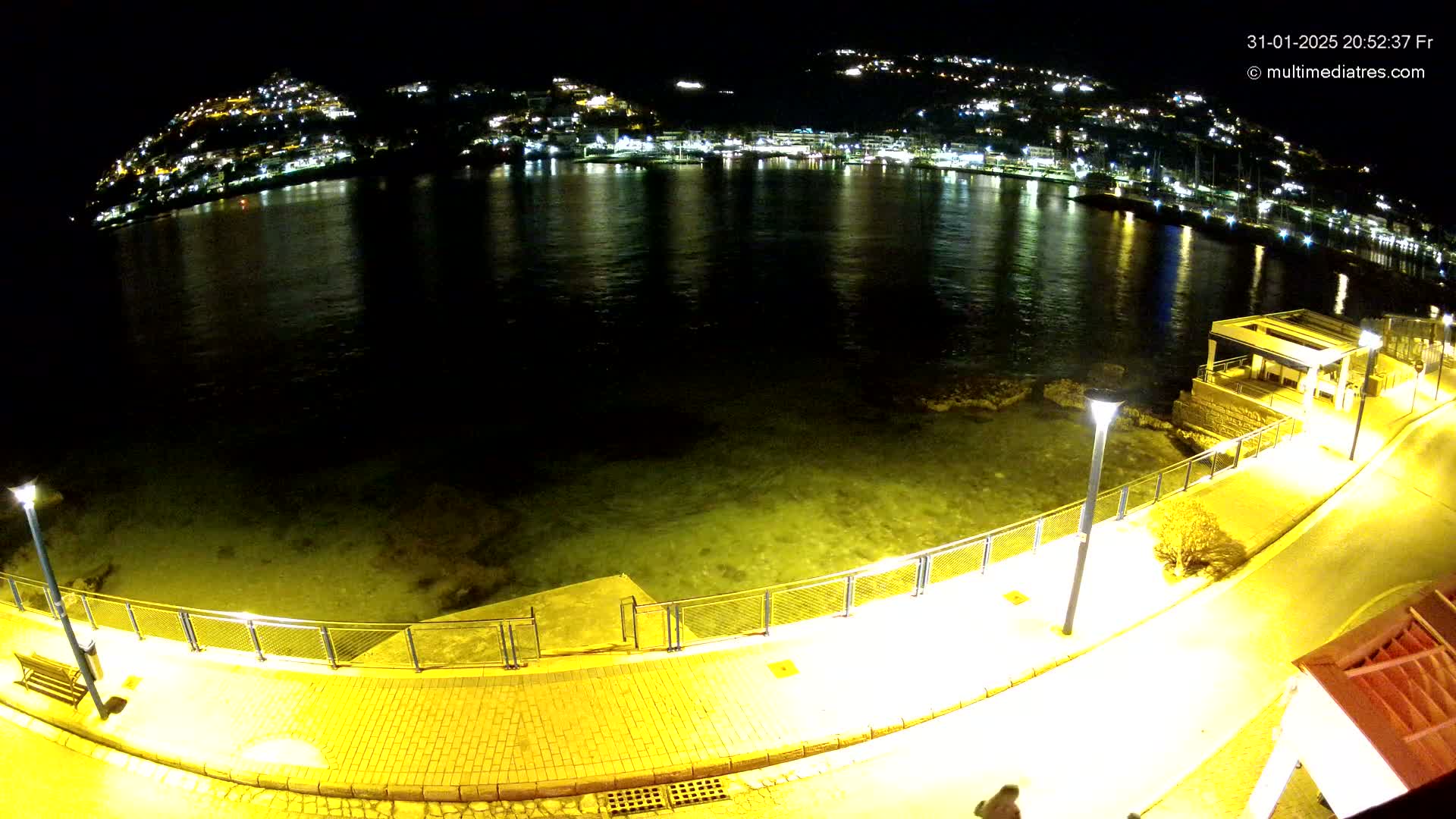 A nighttime, waterfront view shows a calm body of water reflecting the lights of a town nestled on a hillside across the water, with a paved walkway and lights along the shore in the foreground.