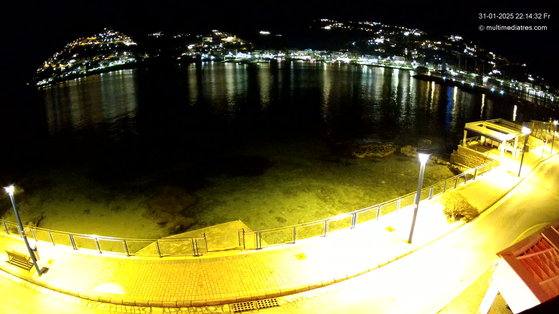 A nighttime view of a calm bay reflecting the lights of a hillside town, seen from a paved waterfront path illuminated by lampposts.