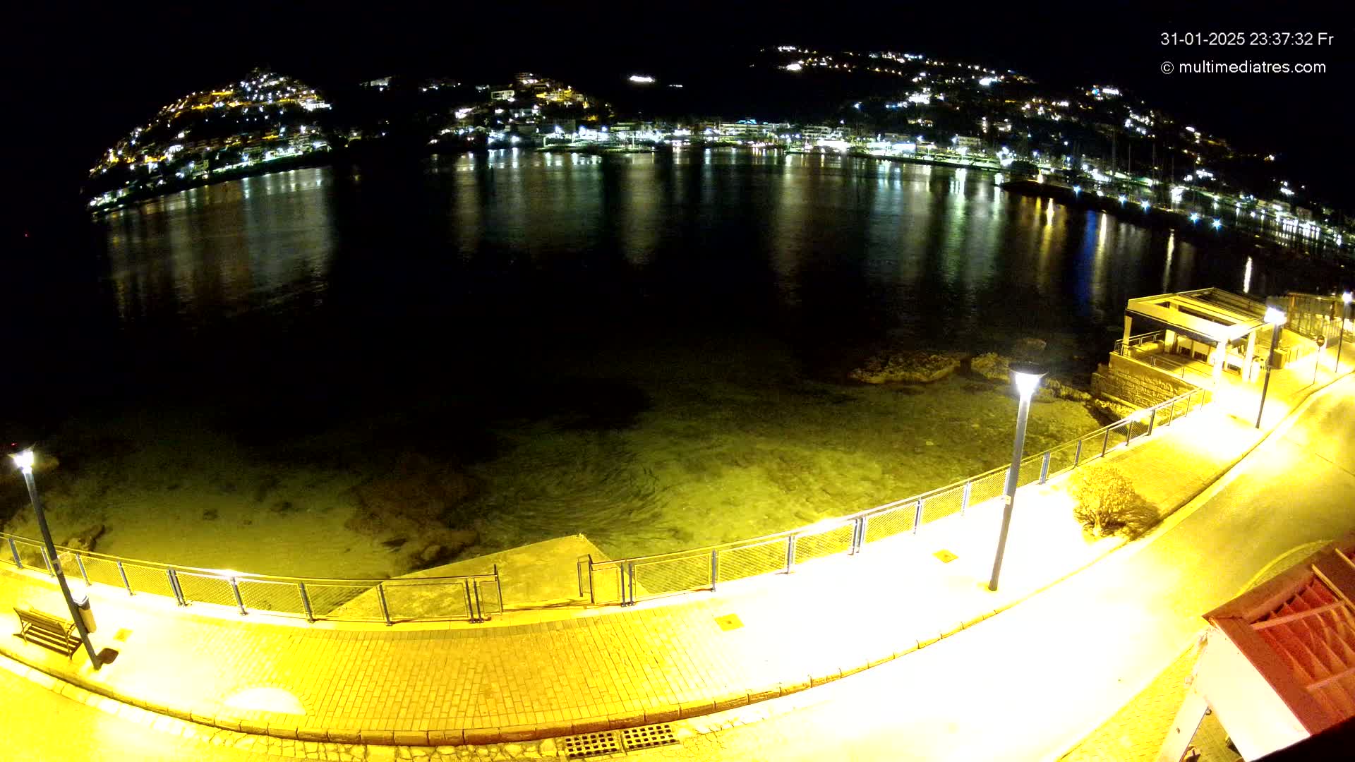 A nighttime view of a calm, shallow body of water reflecting the lights of a town built on a hillside, with a paved walkway and lamps lining the shoreline.