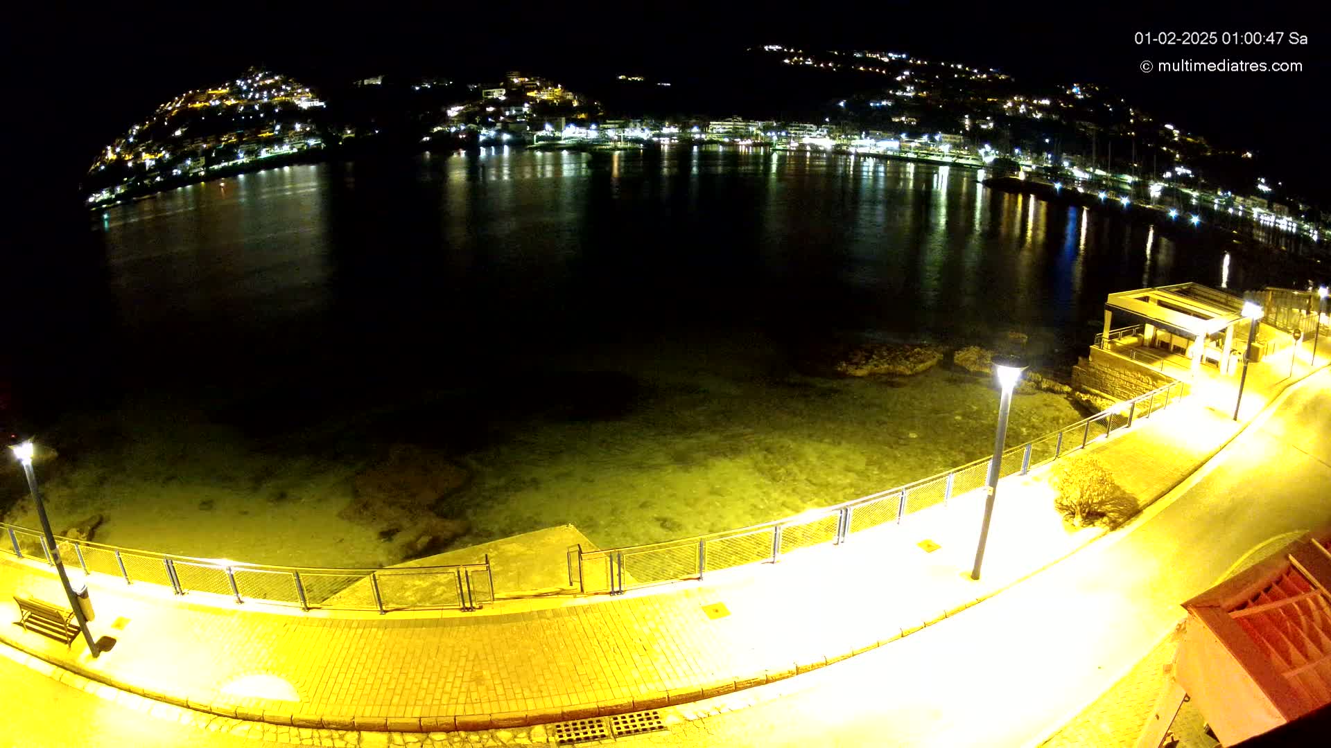 A nighttime view of a calm bay with a town's lights illuminating the water, seen from a paved walkway with lights along a seawall.