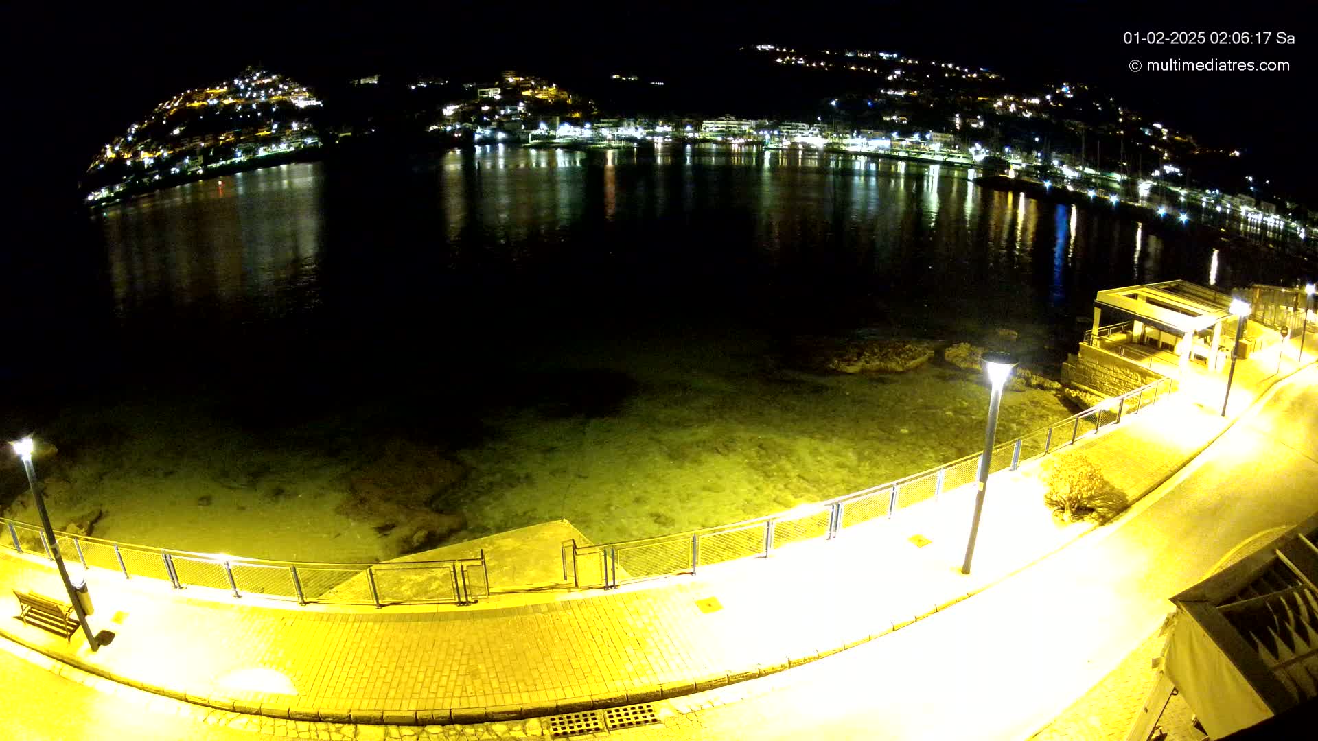 A nighttime, high-angle view shows a calm body of water reflecting the lights of a hillside town, with a paved walkway and railing along the shore illuminated by lampposts.