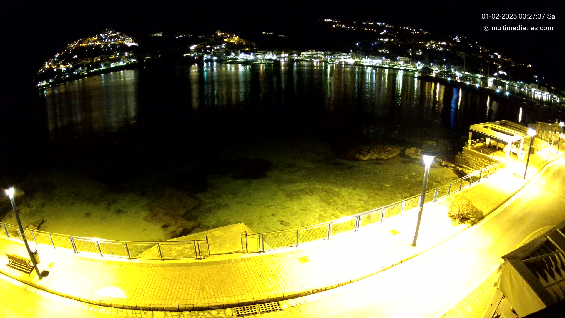 A nighttime, aerial view shows a calm body of water reflecting the lights of a town built on a hillside, with a paved walkway and lights along the waterfront.