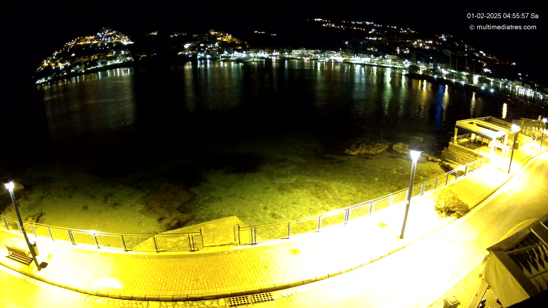 A nighttime view of a calm body of water reflecting the lights of a hilltop town, with a paved walkway and metal fencing bordering the water's edge.