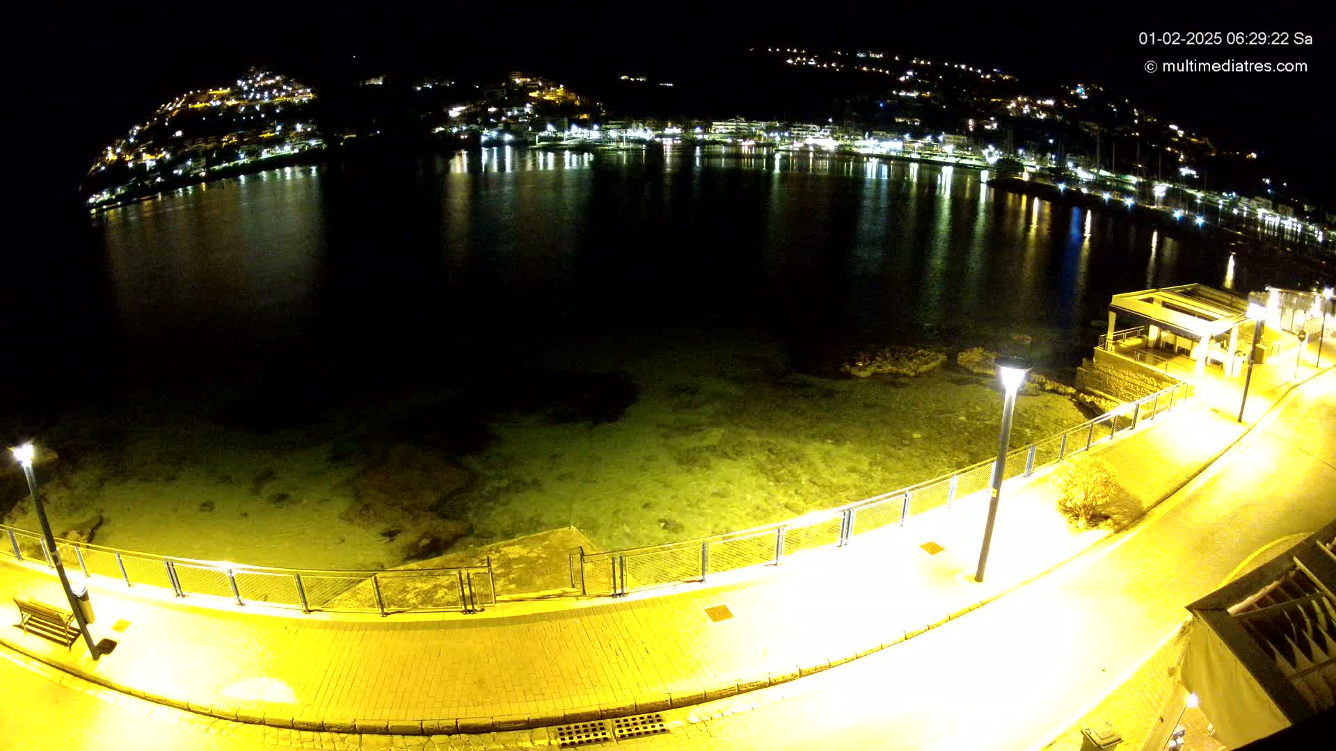 A nighttime, high-angle view shows a calm, shallow body of water reflecting the lights of a hillside town, with a brightly lit paved walkway bordering the water.