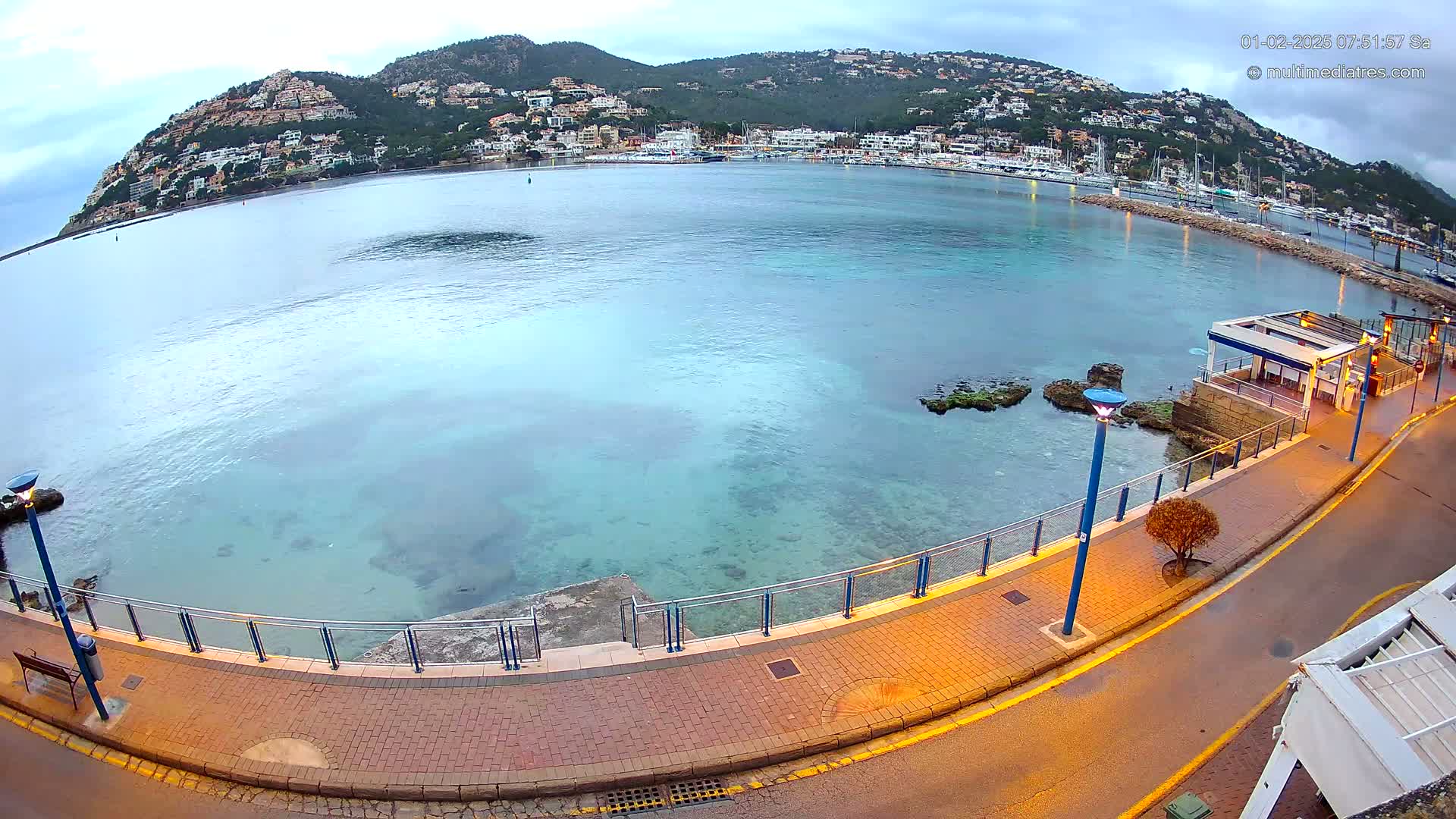 A calm, clear, light-blue bay with a town nestled on a hillside in the background, viewed from a paved walkway next to the water under an overcast sky.
