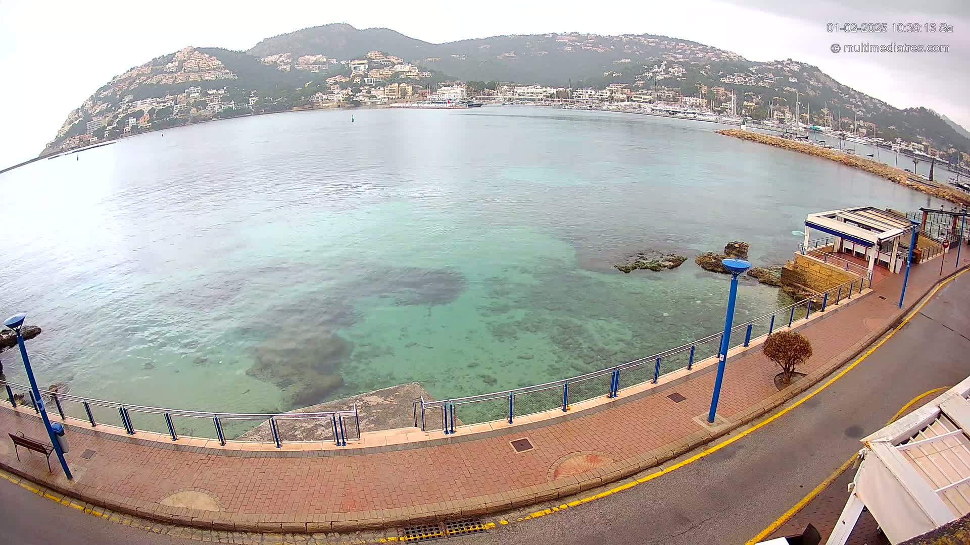 A paved waterfront walkway overlooks a calm, clear turquoise bay with a hillside town in the background under an overcast sky.