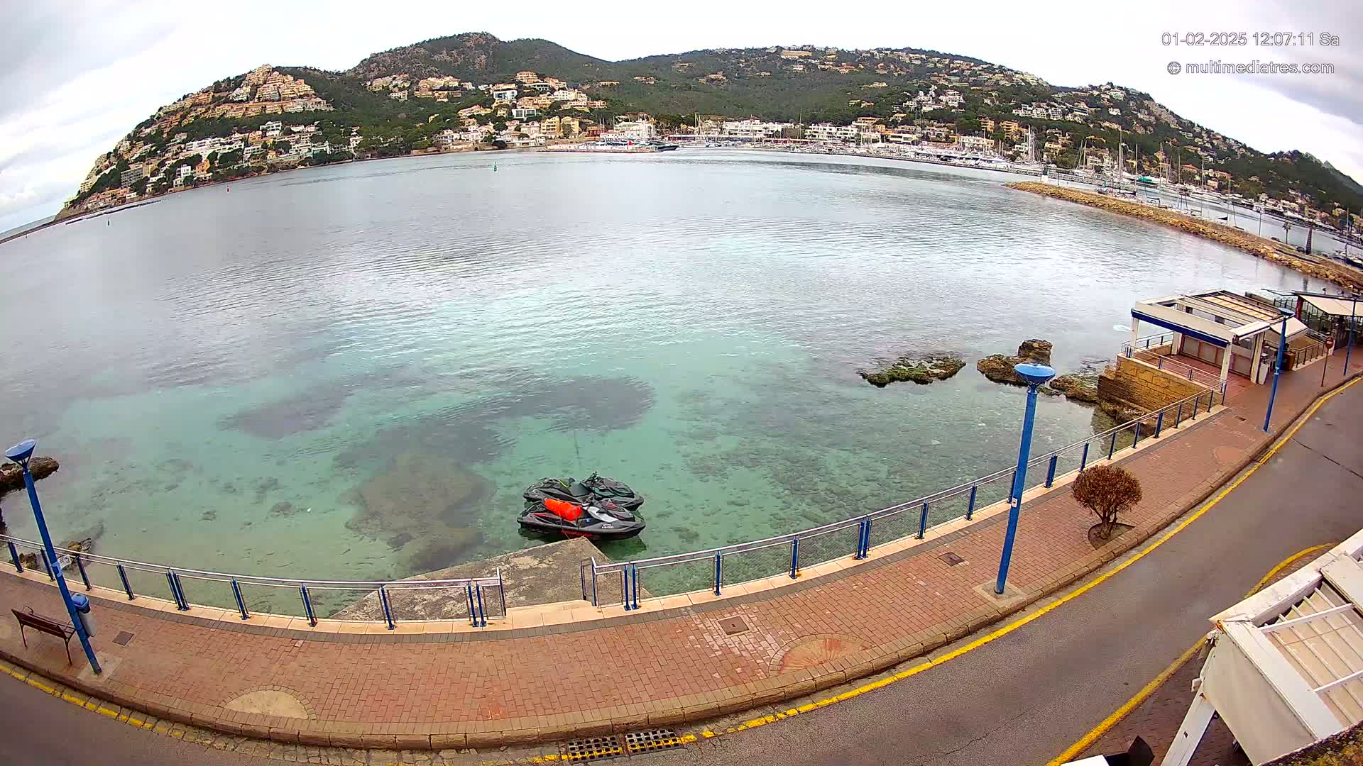 A waterfront paved walkway overlooks a calm bay with clear, shallow water and two jet skis docked near a small building, all under an overcast sky.