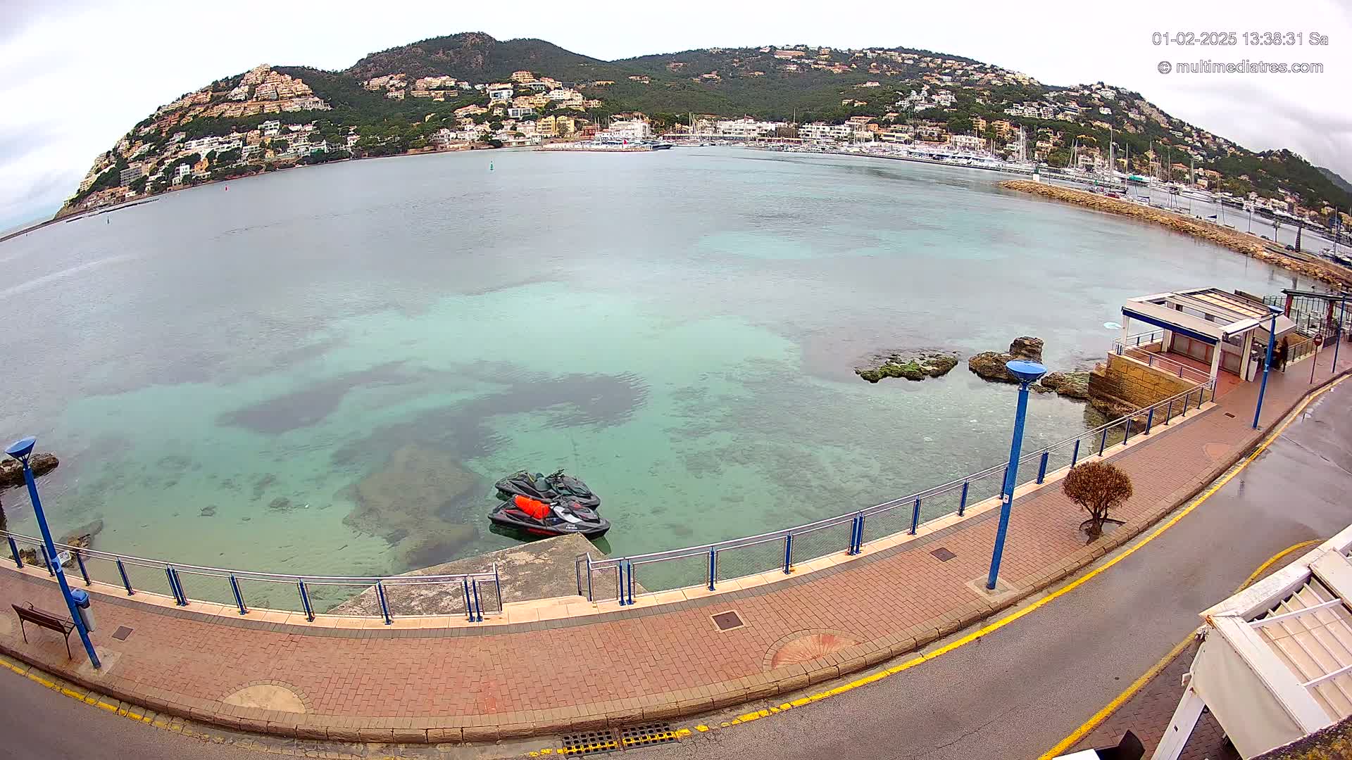 A waterfront paved walkway overlooks a calm, clear teal sea with two jet skis docked near a small building, all situated in front of a hillside town under an overcast sky.