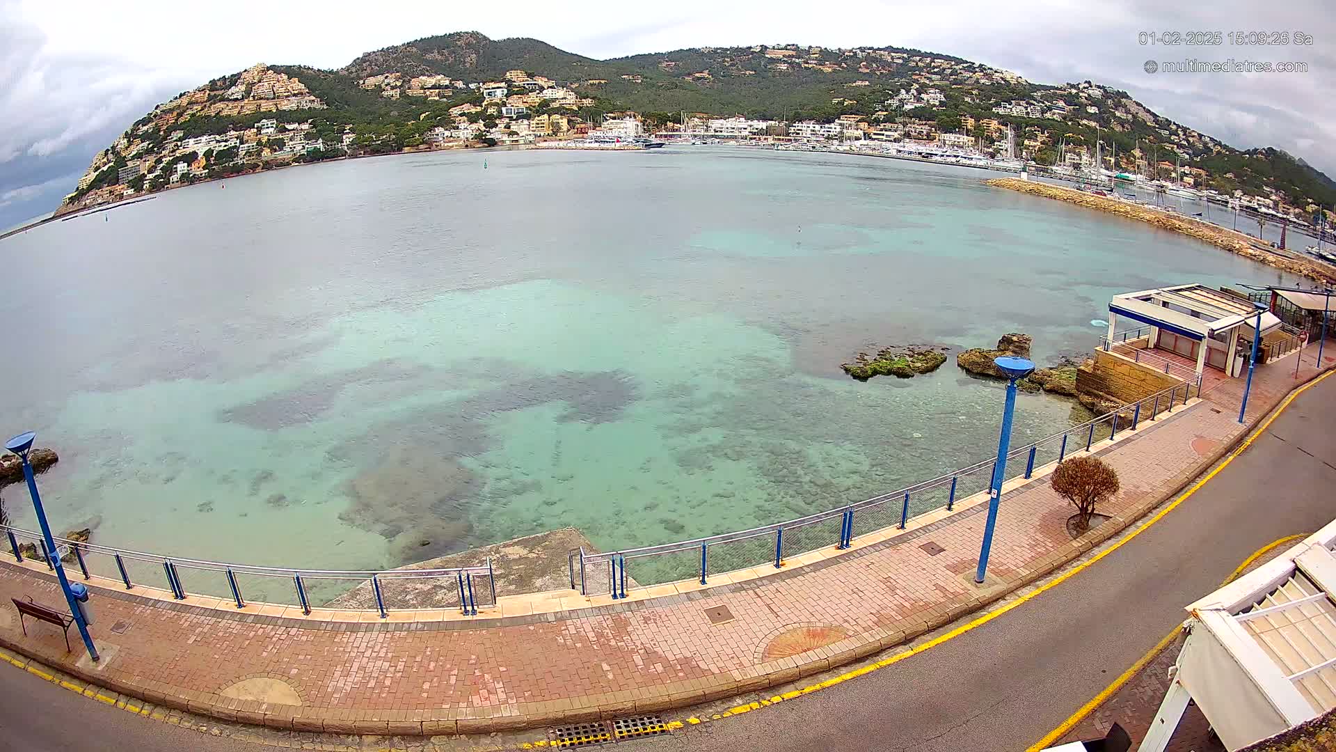 A paved walkway overlooks a calm, clear, teal-colored bay filled with rocky outcrops, bordered by a hillside covered in buildings under an overcast sky.