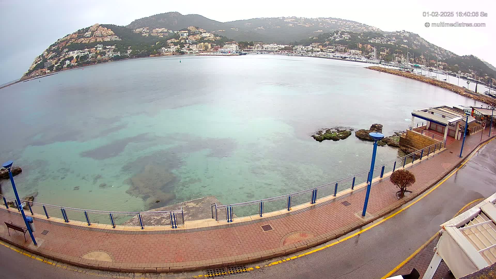A paved walkway curves alongside a calm, clear turquoise bay, overlooking a hillside town under an overcast sky.