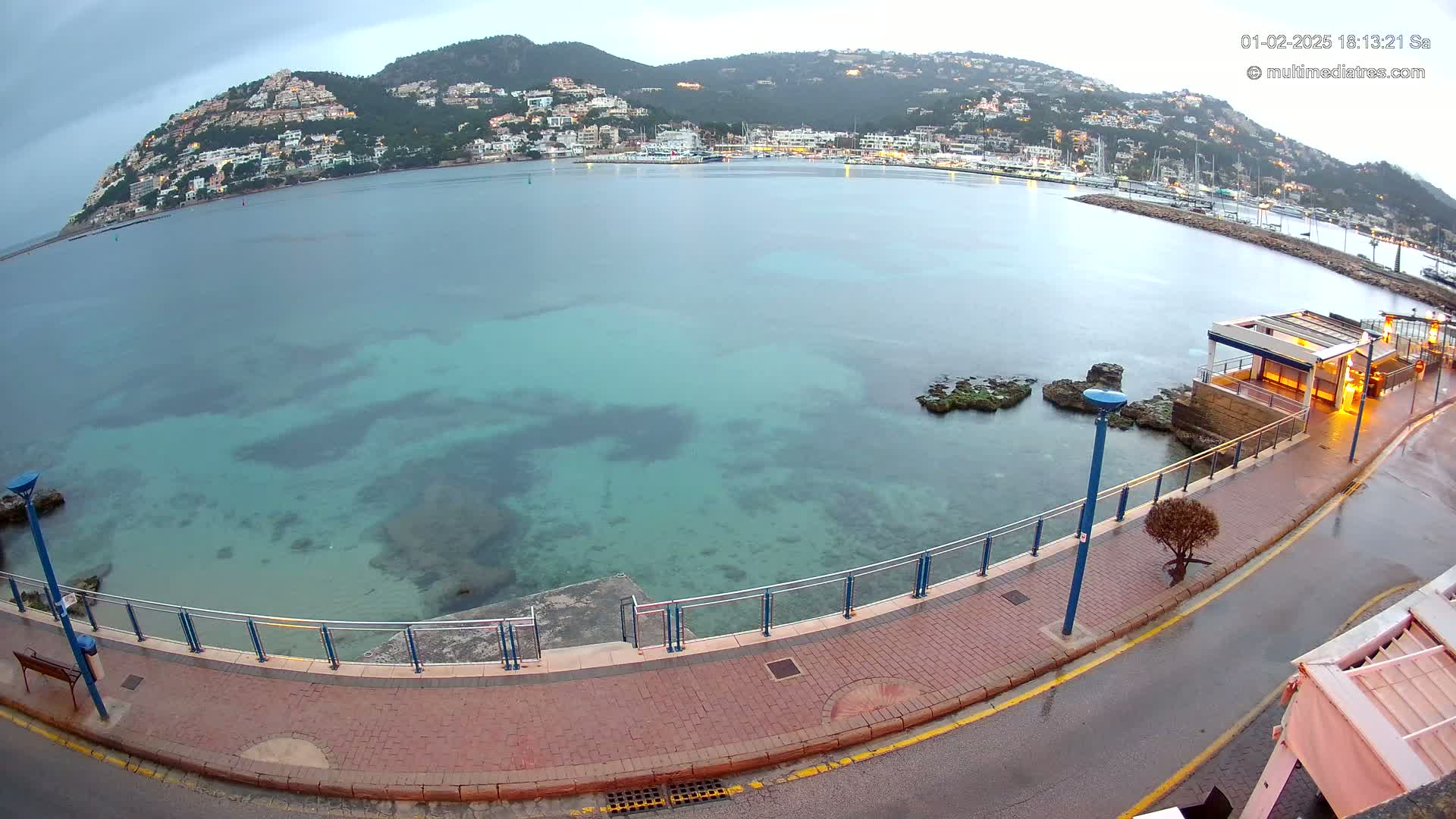 A coastal town is nestled on a hill overlooking a calm, clear turquoise bay under an overcast sky.