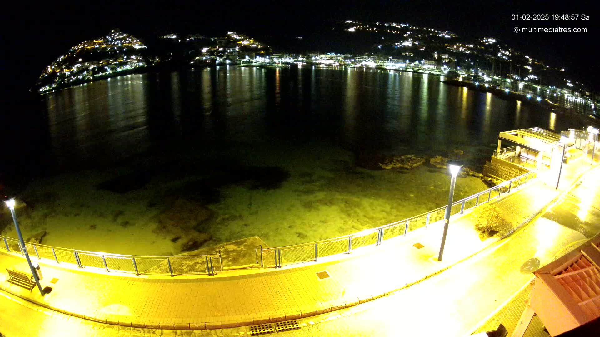 A nighttime view of a calm, shallow body of water reflecting the lights of a hillside town, seen from an illuminated walkway.