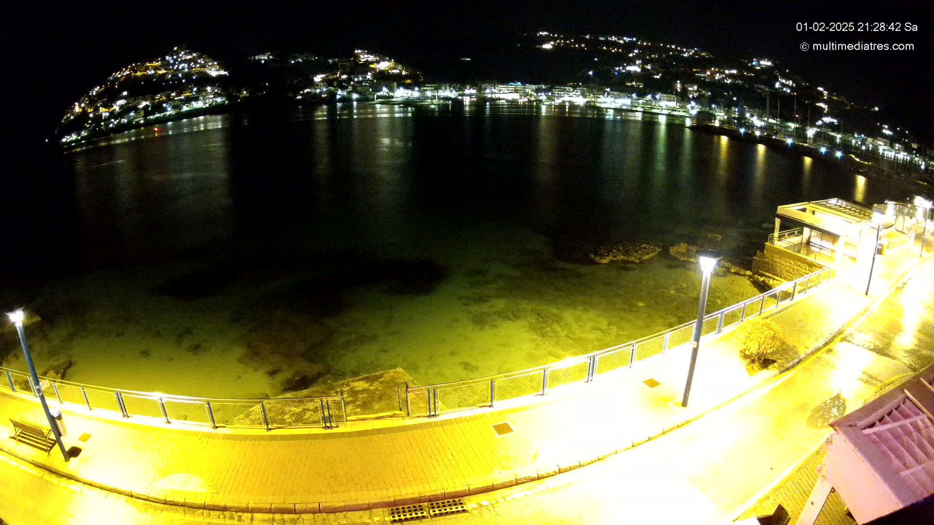 A nighttime, illuminated seaside walkway overlooks calm water and a town on a hill across the bay.