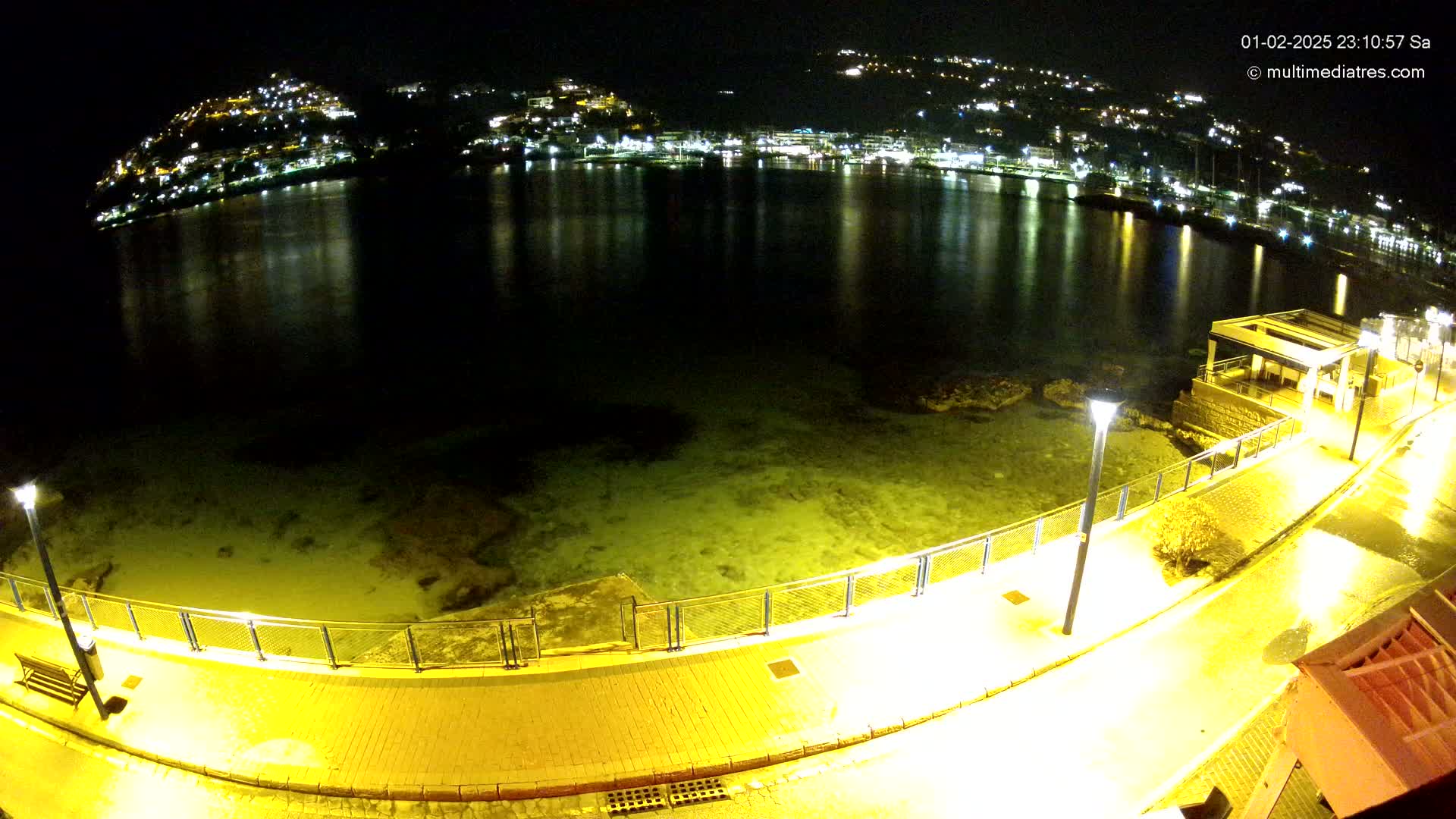 A nighttime view of a calm, illuminated body of water reflecting the lights of a town on a hill, seen from a waterfront walkway with lamps.