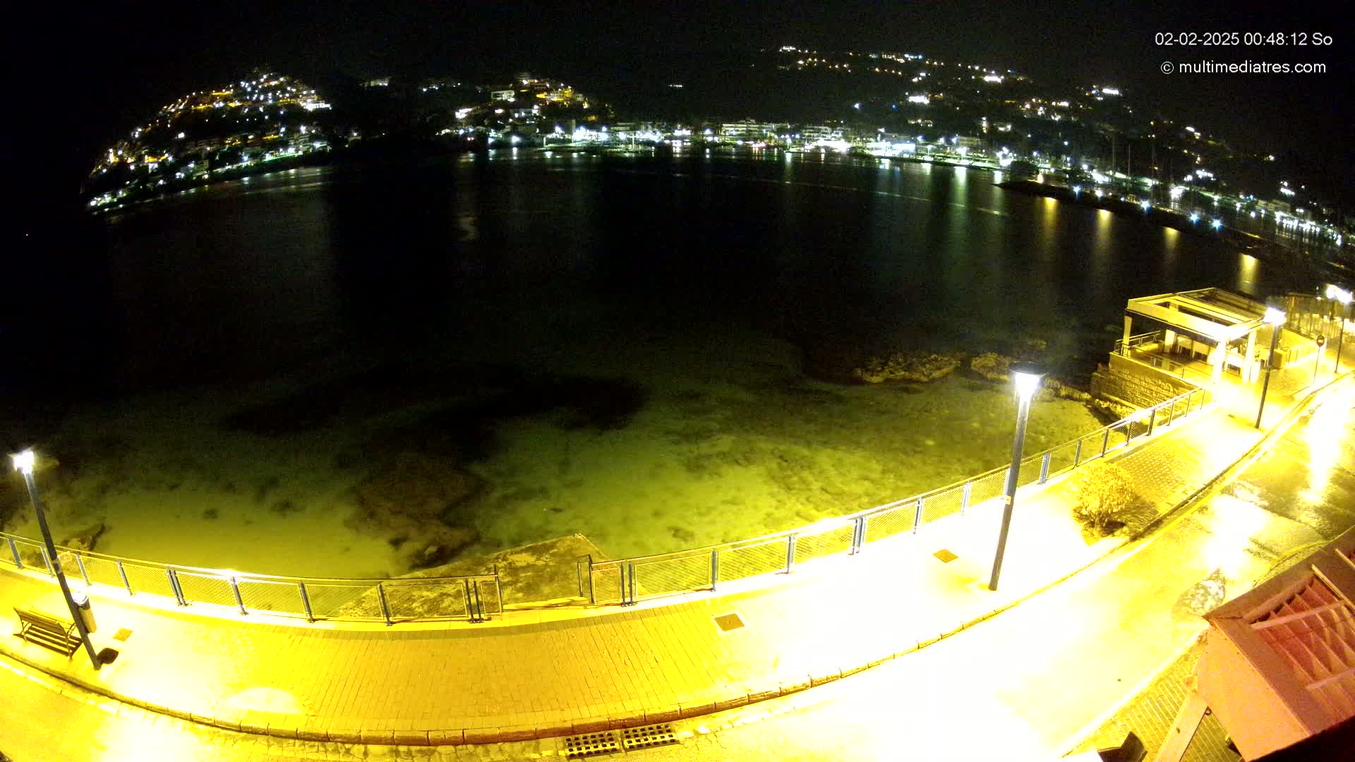 A nighttime, illuminated seaside promenade curves around a calm, shallow body of water, overlooking a town speckled with lights on a hillside.