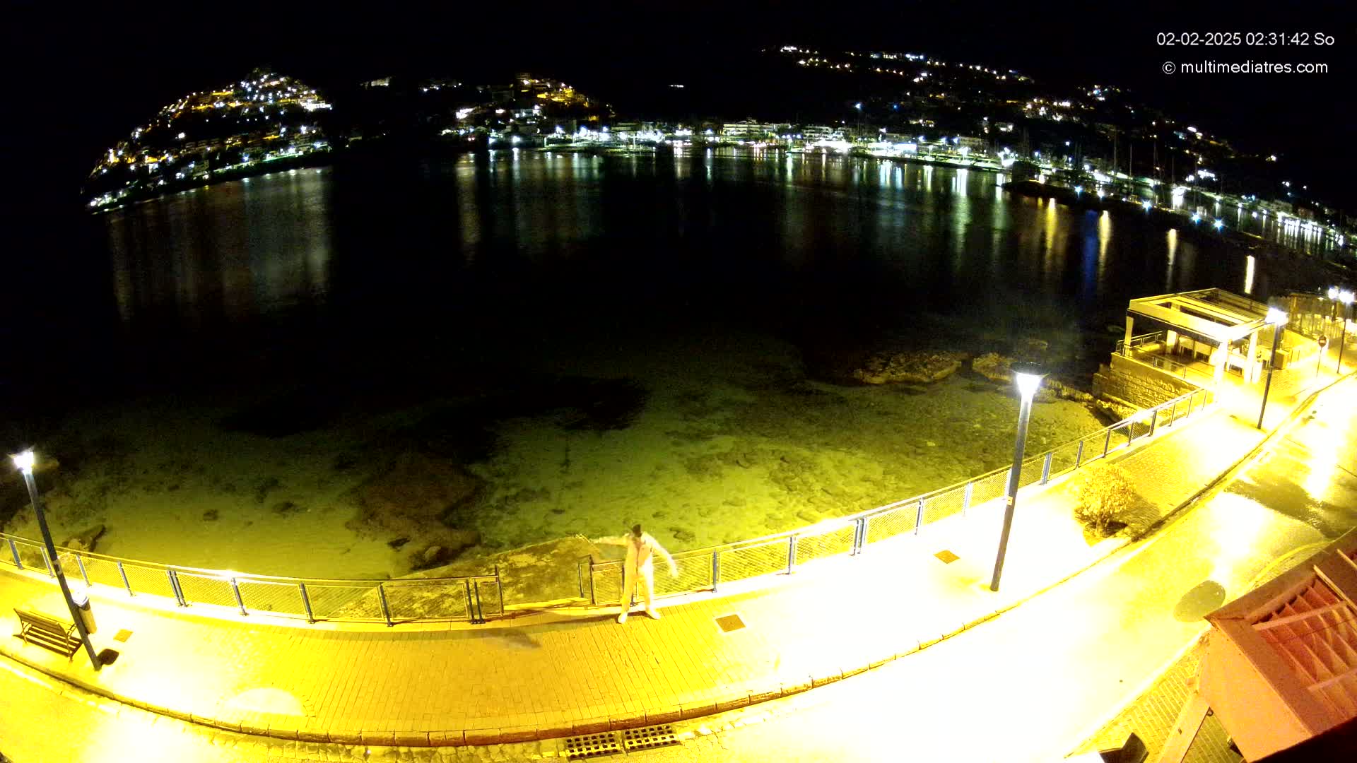 A person stands on a brightly lit walkway at night overlooking a calm body of water reflecting the lights of a town on the hillside beyond.