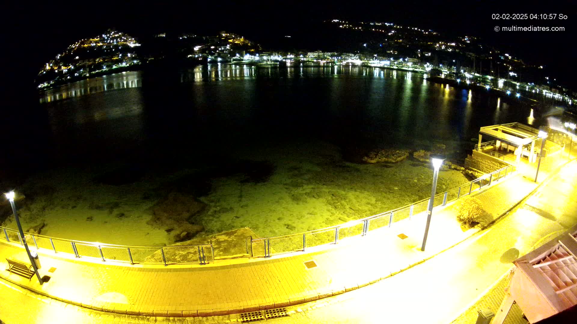 A nighttime view of a calm bay, illuminated by city lights reflected on the water, with a paved walkway and lights along a shoreline.