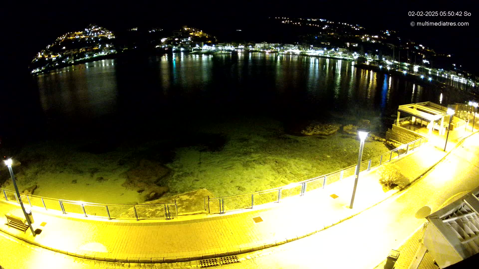 A nighttime view of a calm, shallow body of water next to a lit walkway, with a hilltop town illuminated in the background.
