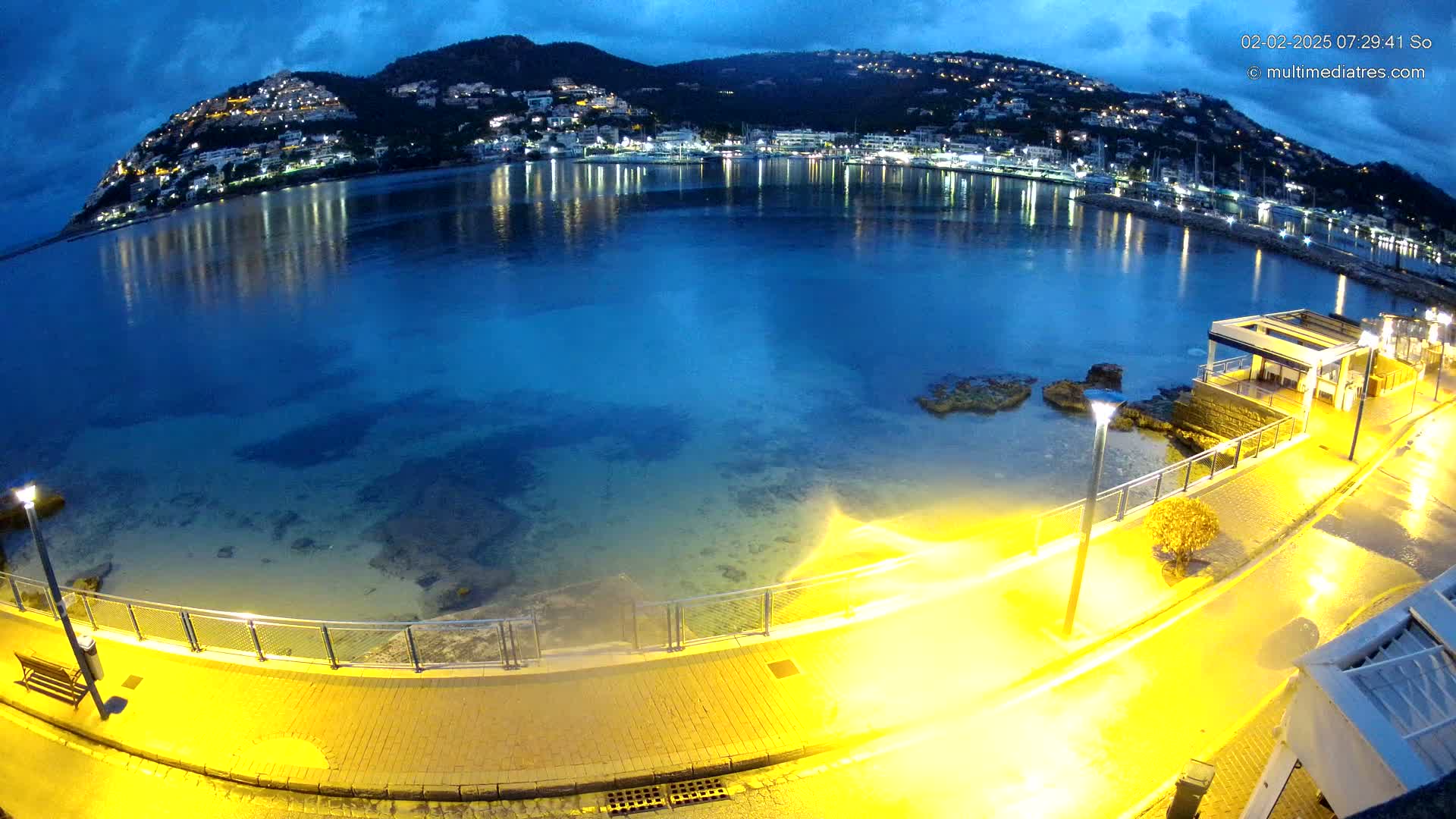 A nighttime view of a calm bay with a hilltop town illuminated against a dark blue, cloudy sky, seen from a waterfront walkway.