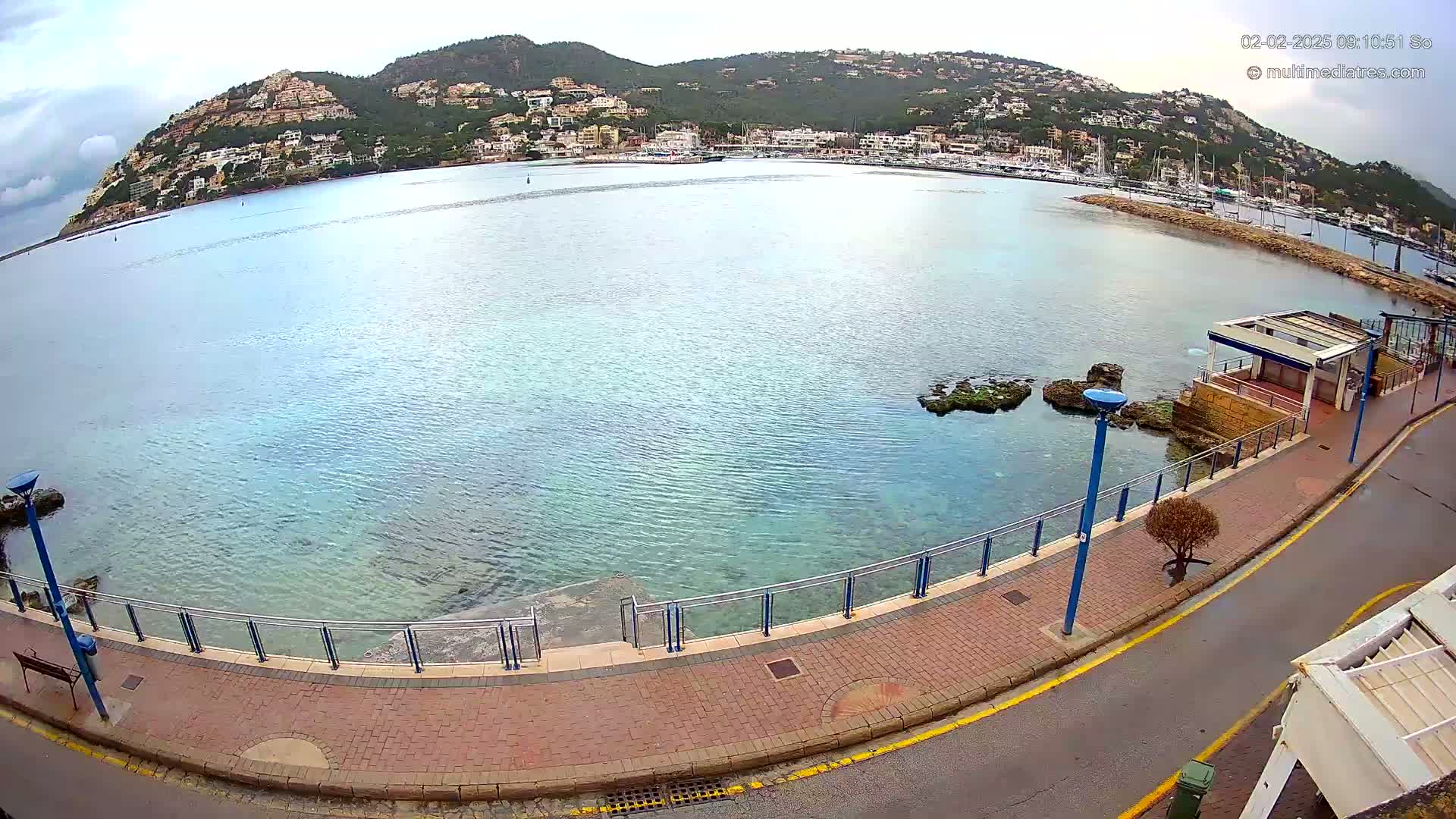 A paved waterfront walkway with a metal railing overlooks a calm, clear bay, with a hill covered in buildings and vegetation in the background under a partly cloudy sky.
