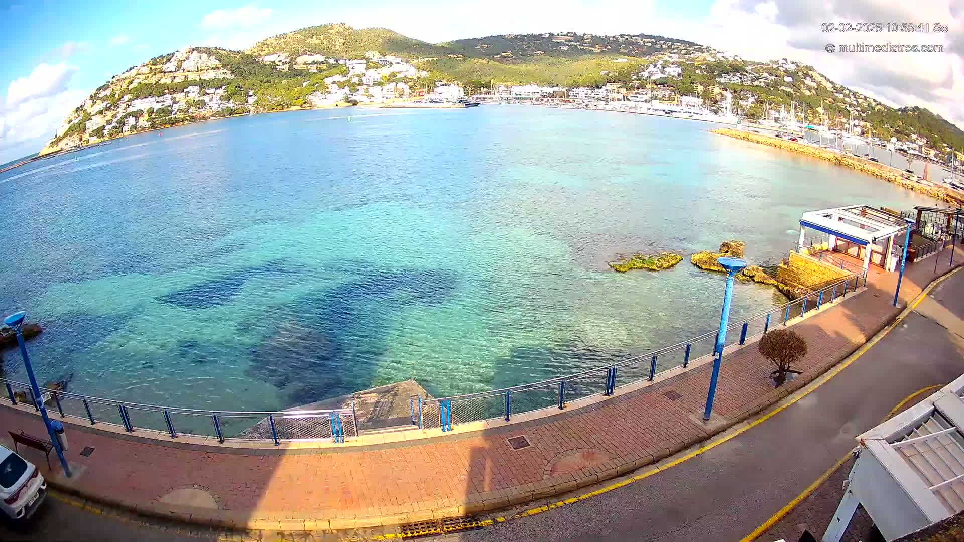 A seaside promenade overlooks clear, shallow turquoise water with a hill dotted with buildings in the background under a mostly sunny sky.